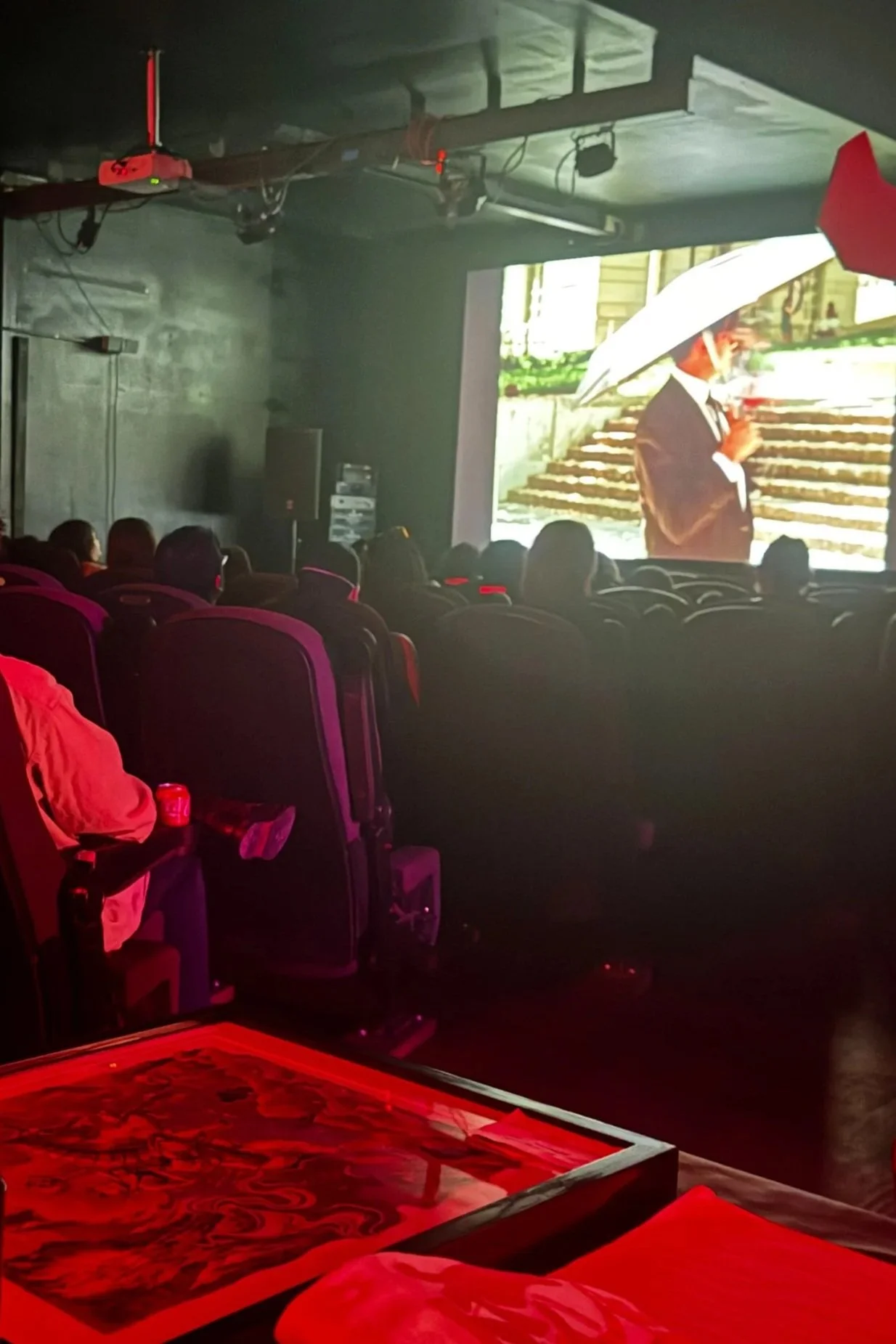 A theater or auditorium showing a large screen with a man in a suit holding an umbrella, outside stairs, and audience members sitting in theater seats.