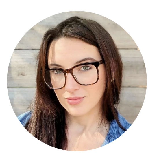 Close-up portrait of a woman with long brown hair, wearing glasses and a denim top, standing in front of a wooden background.