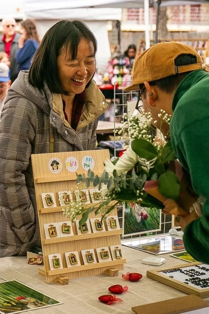 A woman smiling at a vendor selling art pins at a market stall decorated with flowers and various artwork, with other market attendees in the background.
