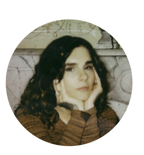 Young woman with dark, curly hair resting her face in her hands, posing indoors against a gray marbled background.