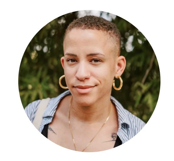 Close-up portrait of a woman with short curly hair, wearing gold hoop earrings, a nose ring, layered necklaces, a striped shirt, and a tattoo on her collarbone, standing outdoors with greenery in the background.