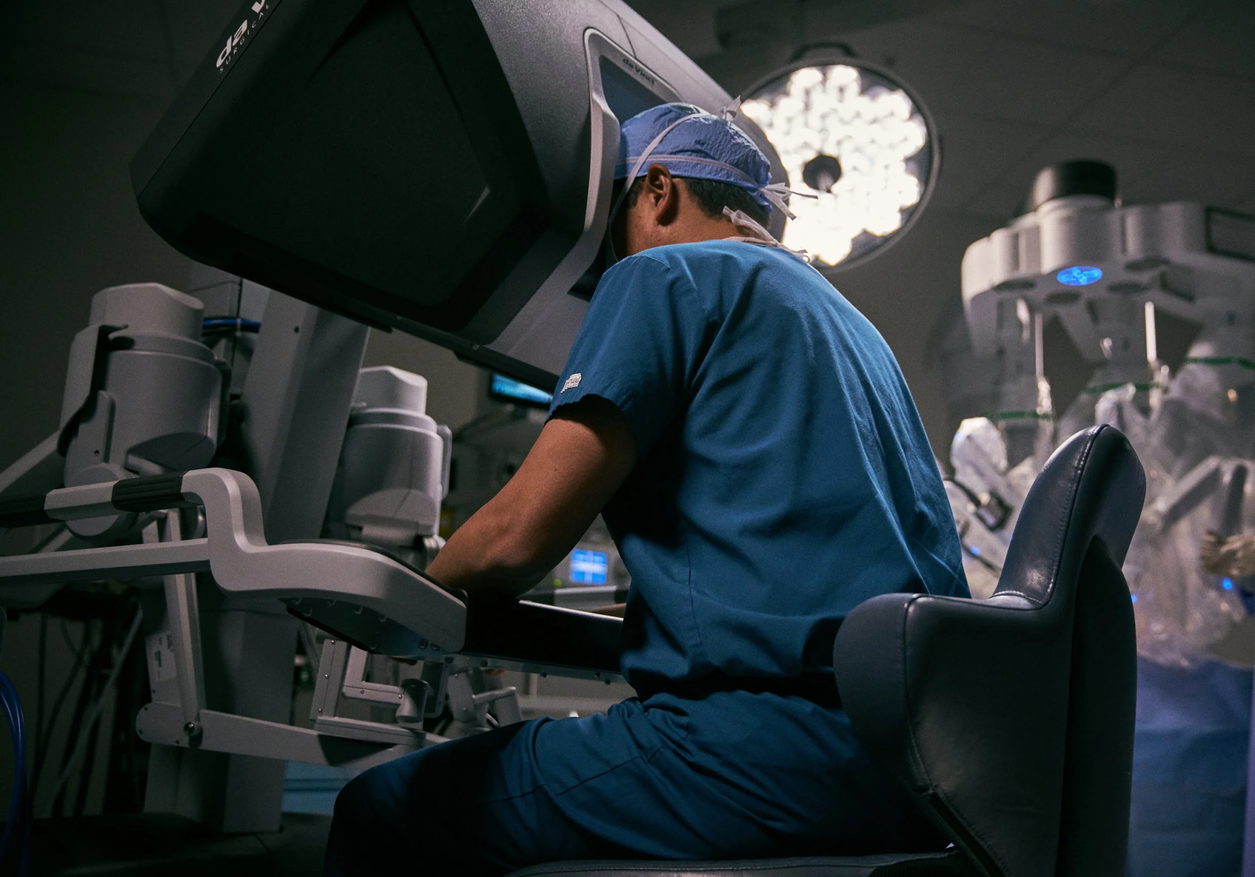 A surgeon in blue scrubs and a surgical cap working in an operating room with medical equipment and overhead lights.