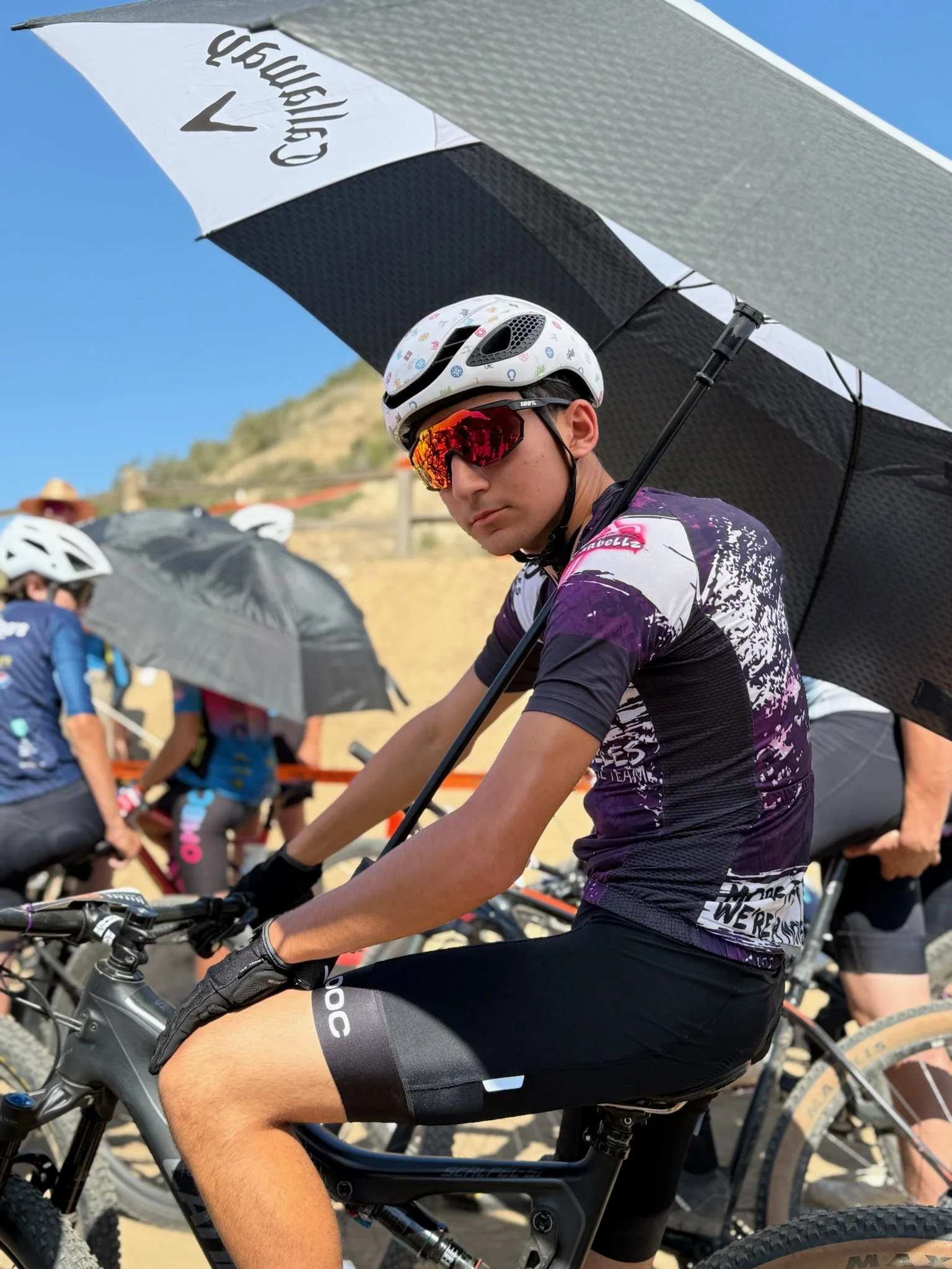 A young man wearing a white cycling helmet with colorful designs and red sports sunglasses, riding a mountain bike during a daytime cycling event, with others and umbrellas in the background.