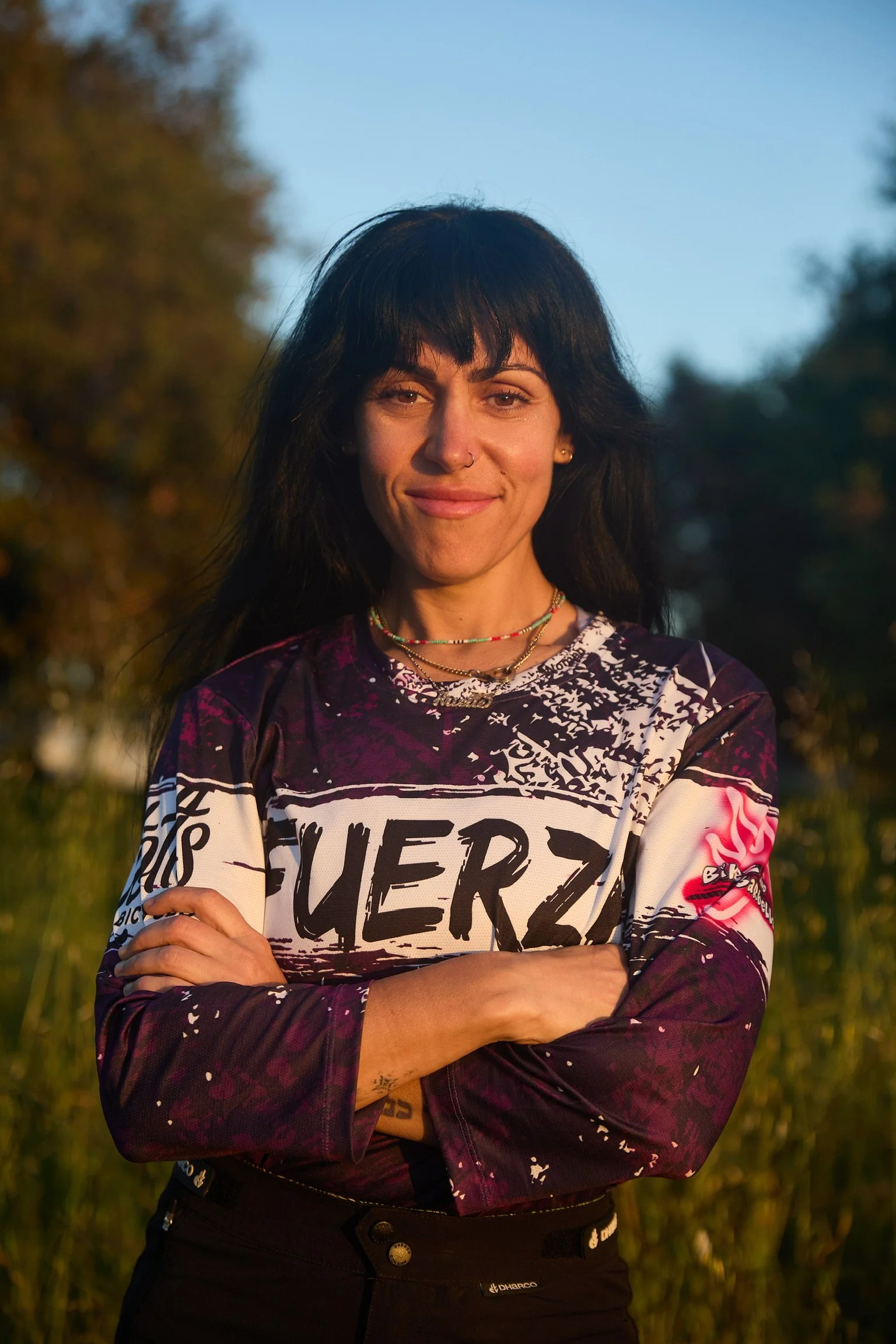 A woman with dark hair, wearing a colorful sports jersey and multiple necklaces, standing outdoors with her arms crossed, smiling at the camera during sunset.