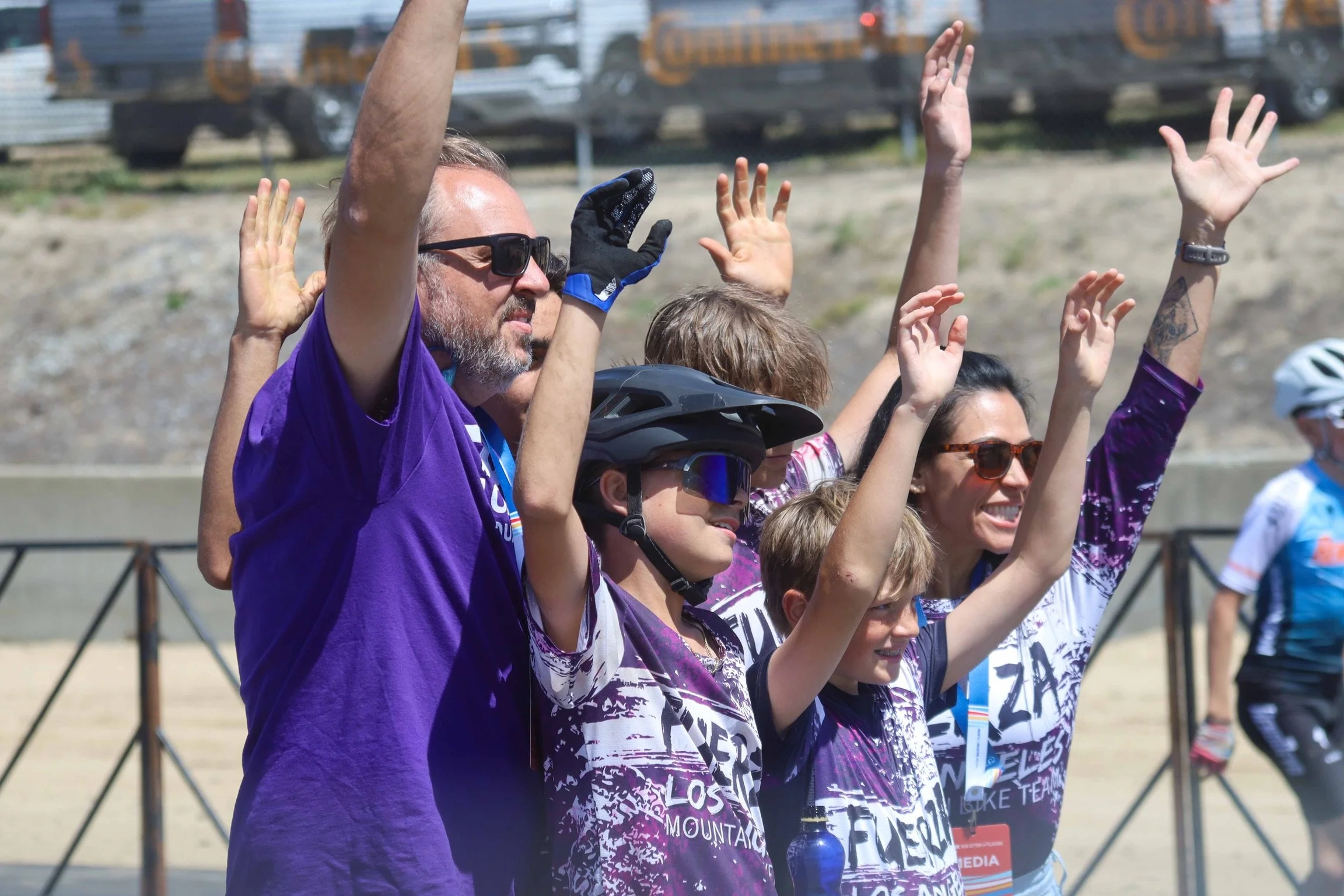 A group of people wearing matching purple and white jerseys, some with helmets and sunglasses, raising their hands in a gesture of celebration or solidarity outdoors on a sunny day.