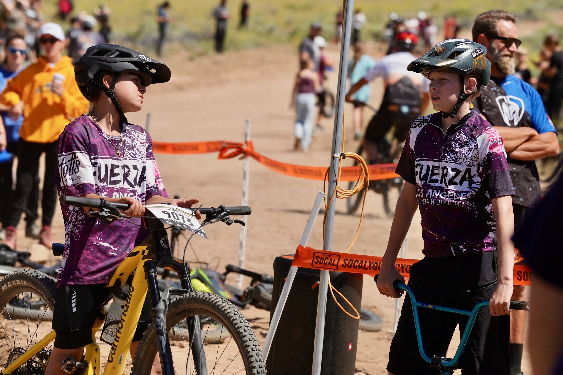 Two young boys wearing helmets and sporty biking jerseys talking near a finishing or starting line at a mountain biking event with spectators in the background.