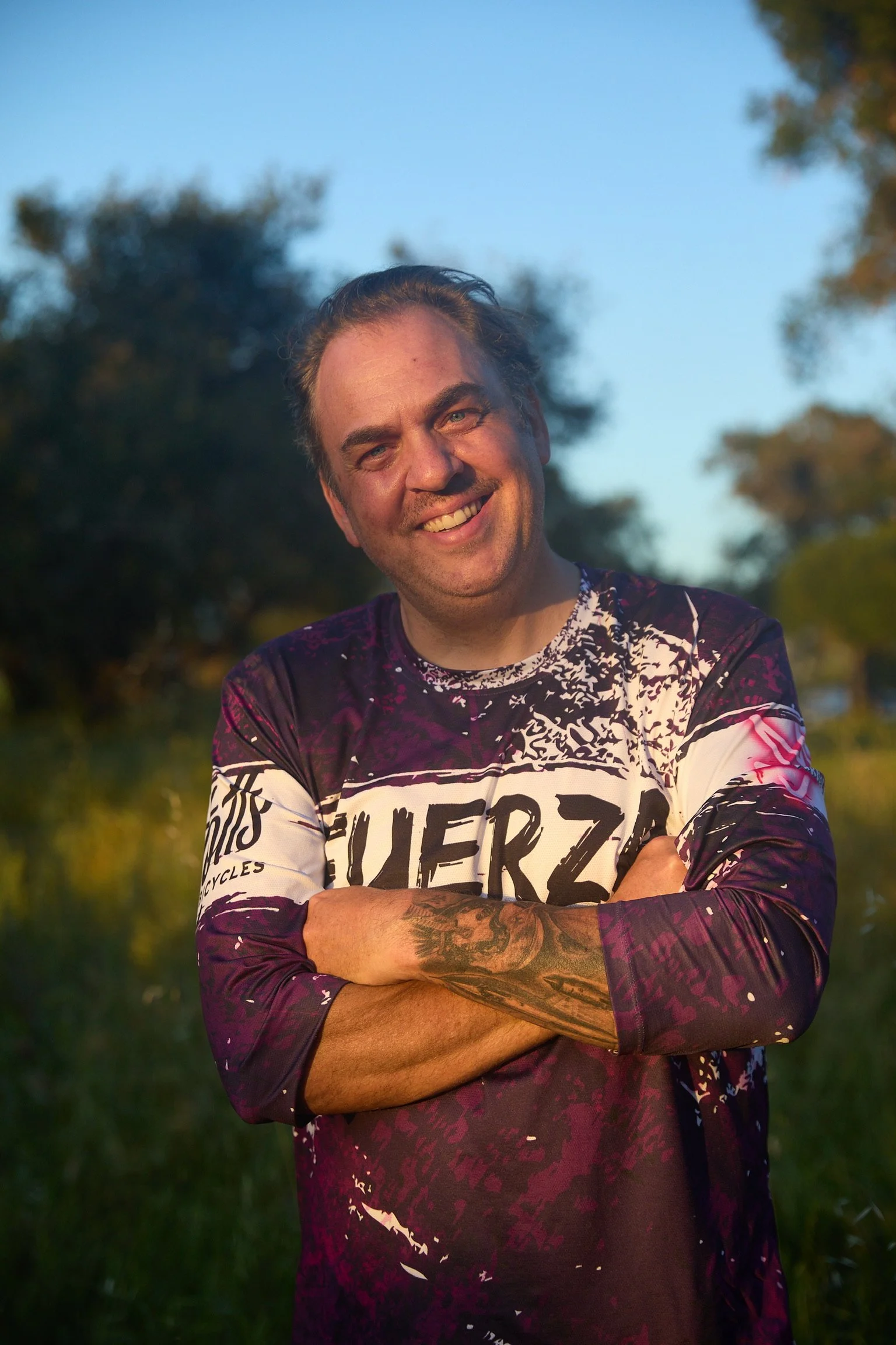 A smiling man with light skin, tattoos on his left forearm, wearing a purple patterned long-sleeve shirt with white text, stands outdoors with his arms crossed. In the background, there's a clear blue sky and blurred trees.