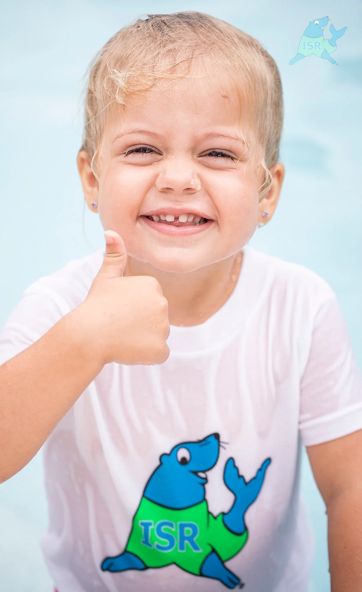 A young girl with blonde hair and a big smile, giving a thumbs-up. She is wearing a white shirt with a blue cartoon bear on it, holding a green and blue hat with the letters "ISR" on it.