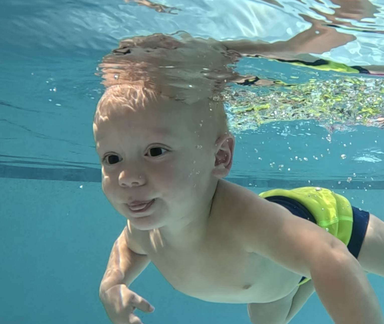 Young child swimming underwater in a pool, with a playful expression and tongue slightly out, wearing yellow and navy swim gear.
