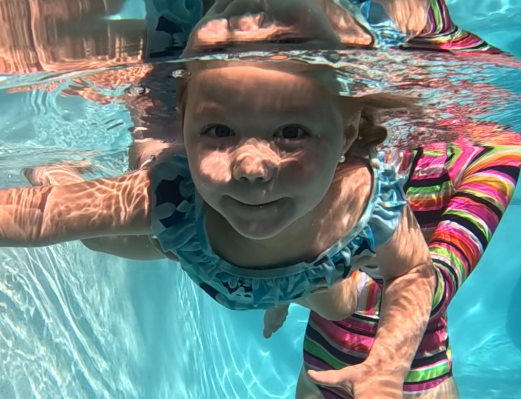 A young girl swimming underwater in a pool, smiling at the camera, wearing a blue swimsuit with ruffled straps.