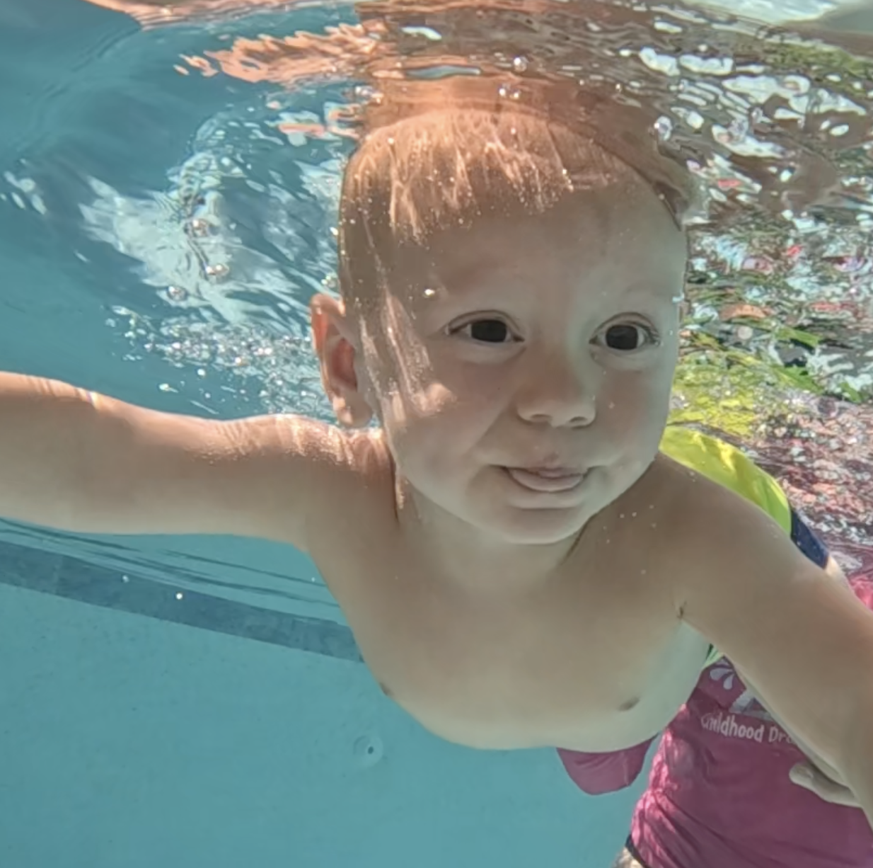 Young child swimming underwater in a pool, smiling at the camera, with water and sunlight reflections around.