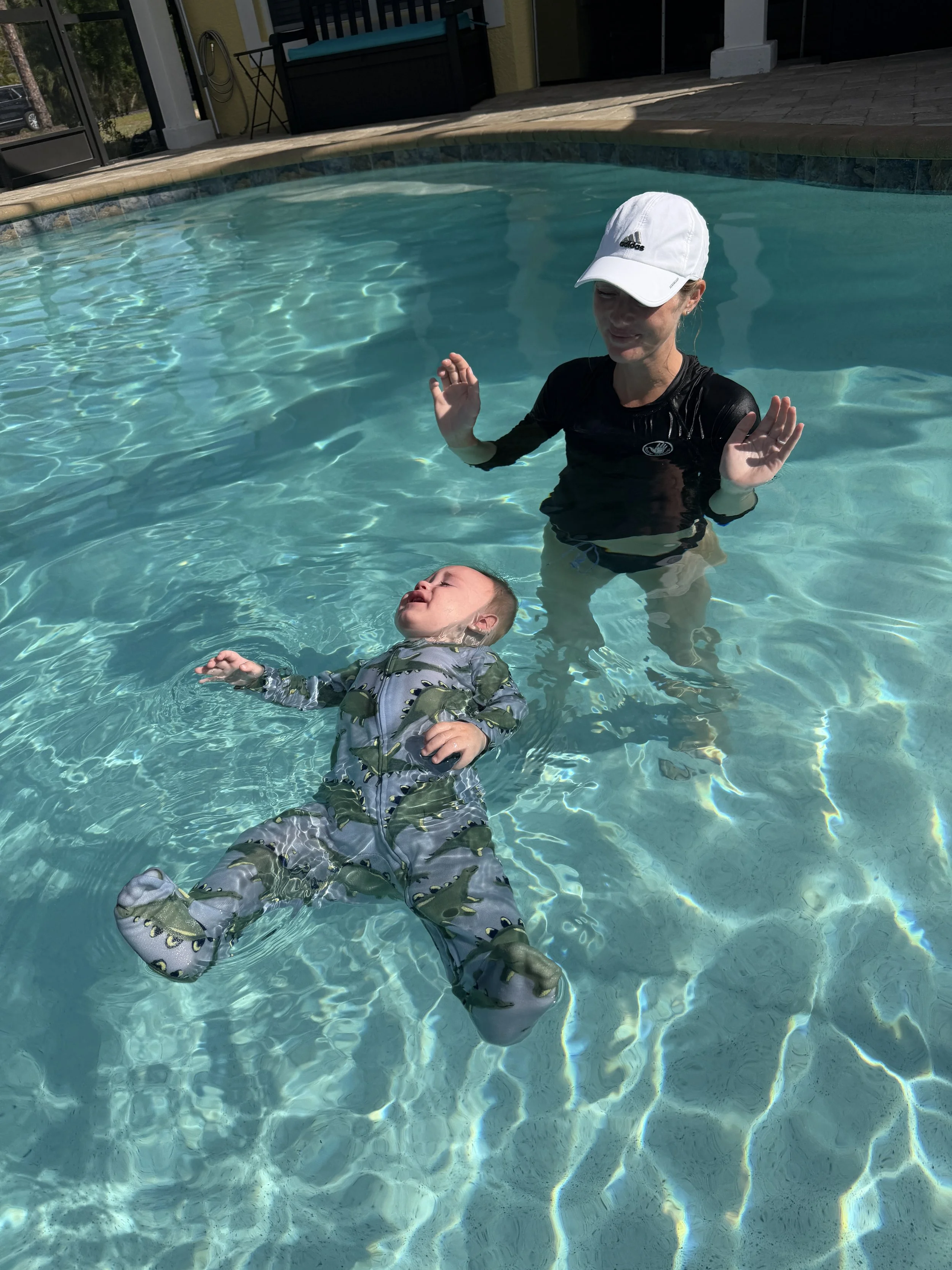 A woman and a child in a swimming pool, with the woman wearing a white cap and black shirt, and the child in a patterned floatation suit, both enjoying a sunny day near a house.