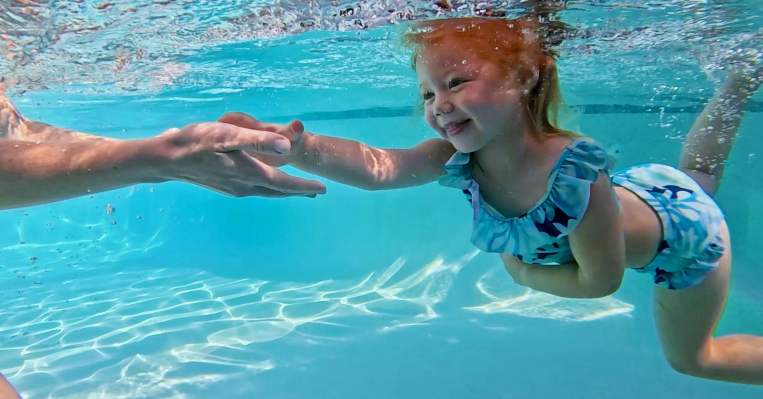 A young girl with blonde hair swimming underwater in a pool, reaching out to high-five an adult's hand, and wearing a blue patterned swimsuit.