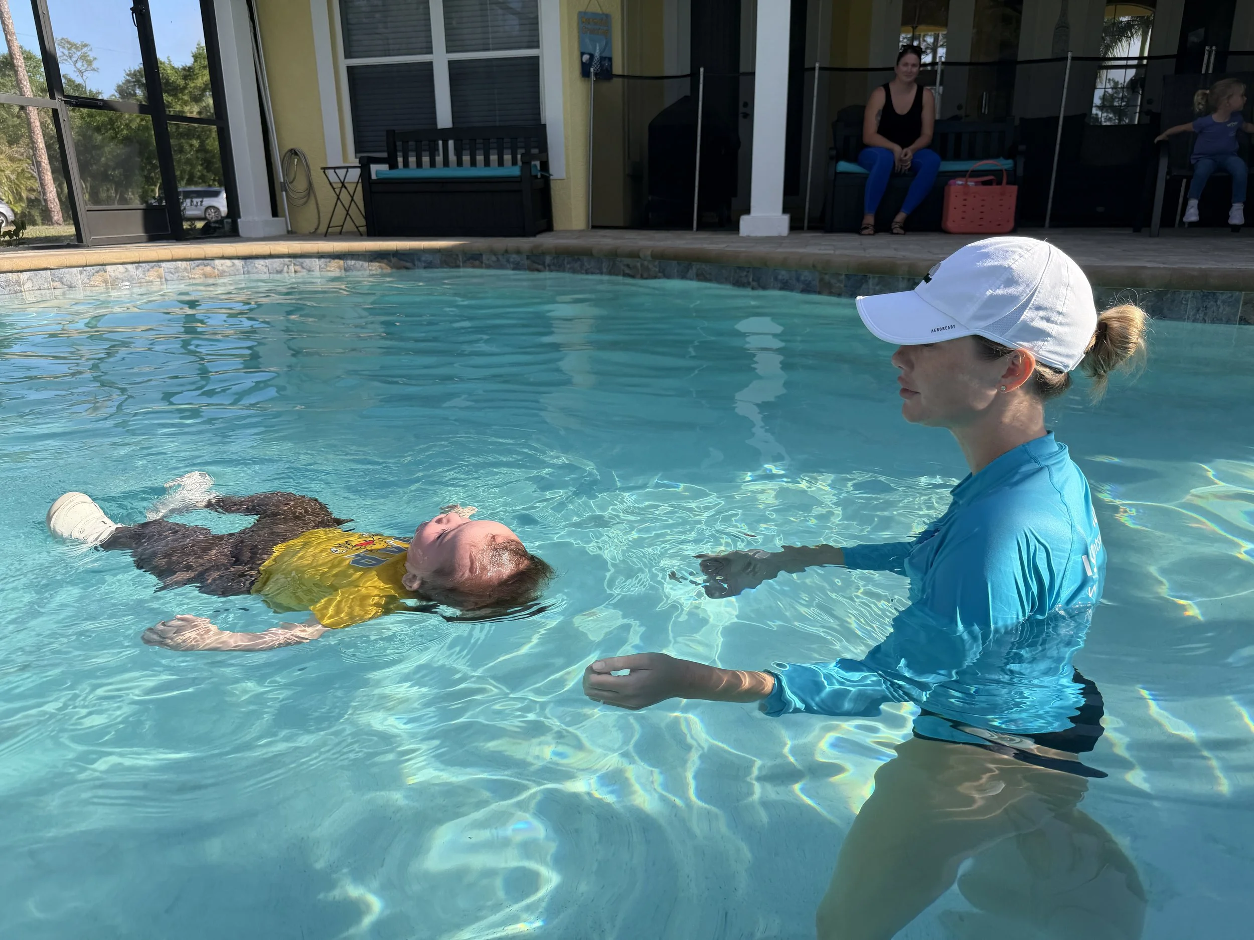 A woman is receiving CPR training in a swimming pool from a female instructor. A child is sitting on a bench in the background outside a house, with trees and a car visible through the windows.