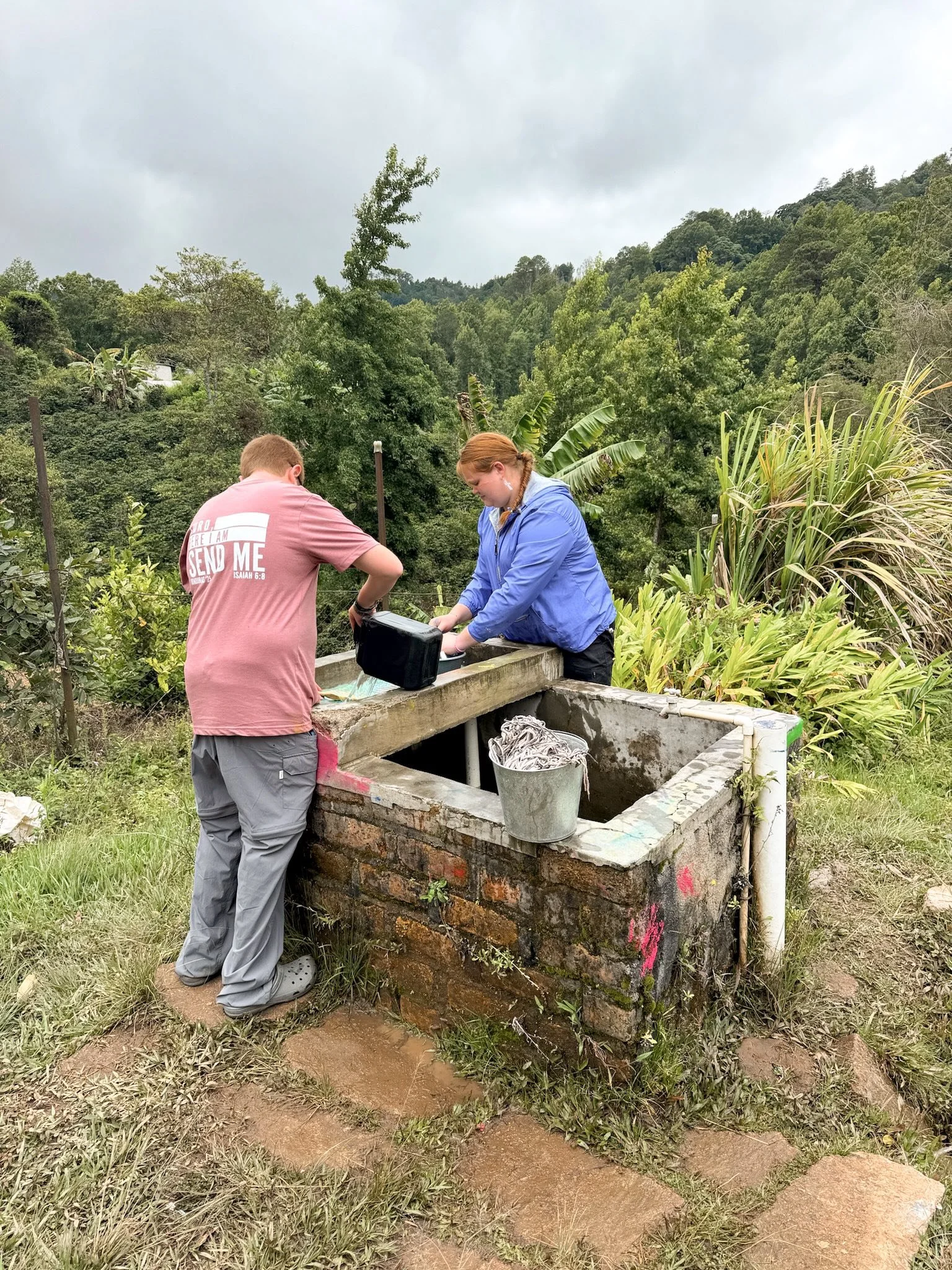 Two people working at an outdoor water faucet with lush green trees and cloudy sky in the background.