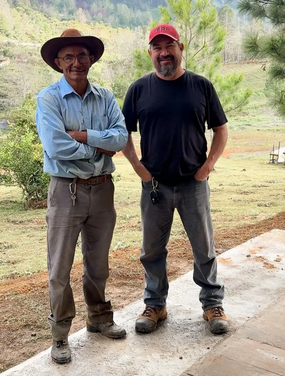 Two men standing outside on a partly cloudy day, one wearing a cowboy hat, glasses, and a light blue shirt with rolled-up sleeves, and the other wearing a red cap, black T-shirt, and gray pants, smiling and posing with hands in pockets.