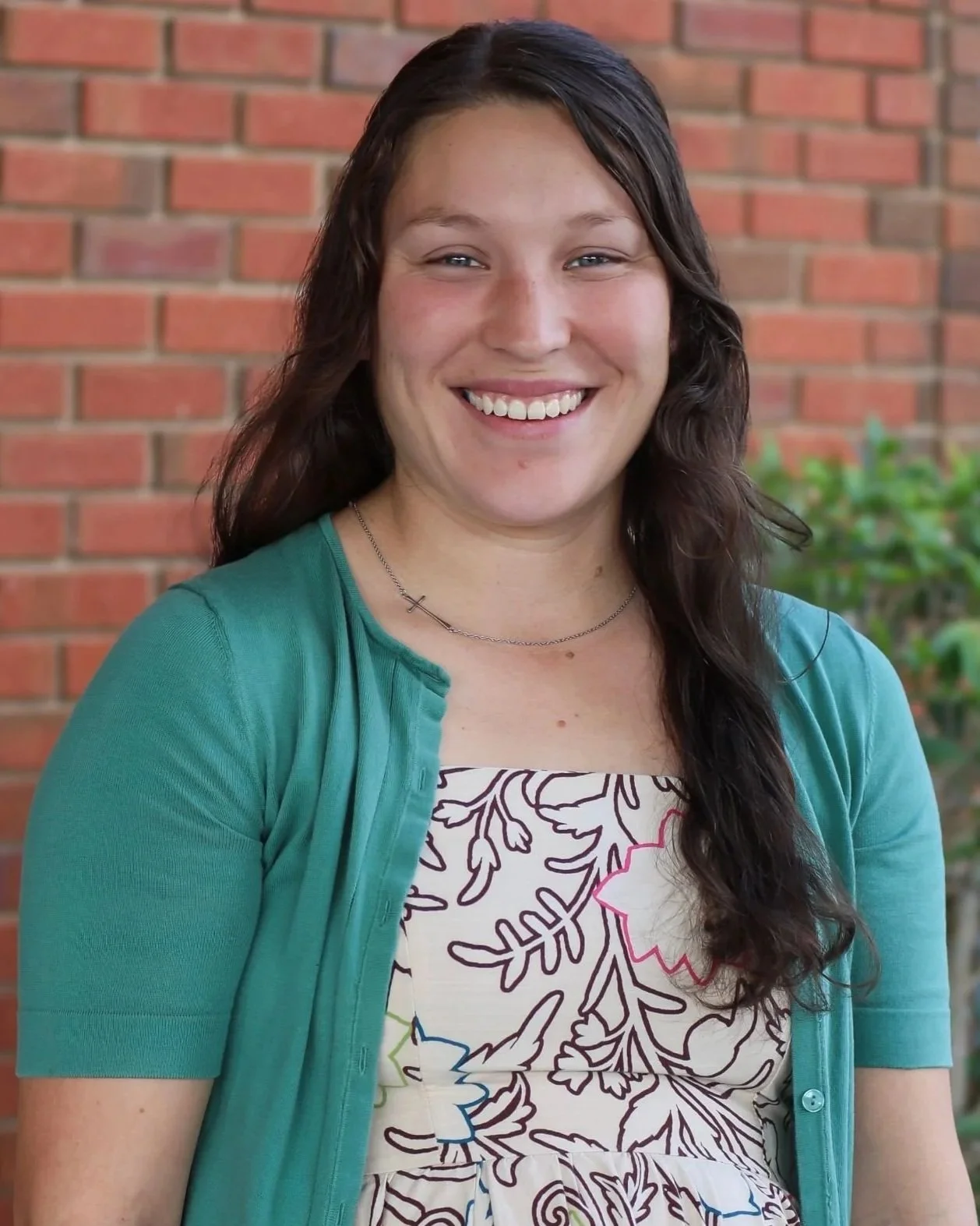 A young woman with long dark hair, smiling, in front of a brick wall and some green foliage, wearing a floral patterned dress and a teal cardigan.