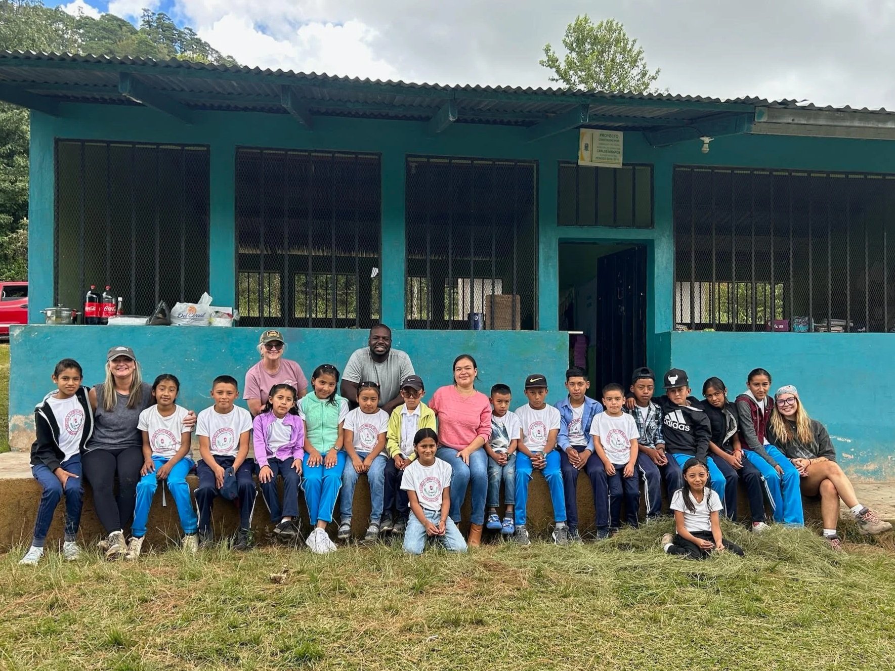 A group of children and adults sitting outdoors in front of a blue building with metal barred windows, surrounded by greenery. Some children are wearing white shirts with a logo, and several are smiling at the camera.