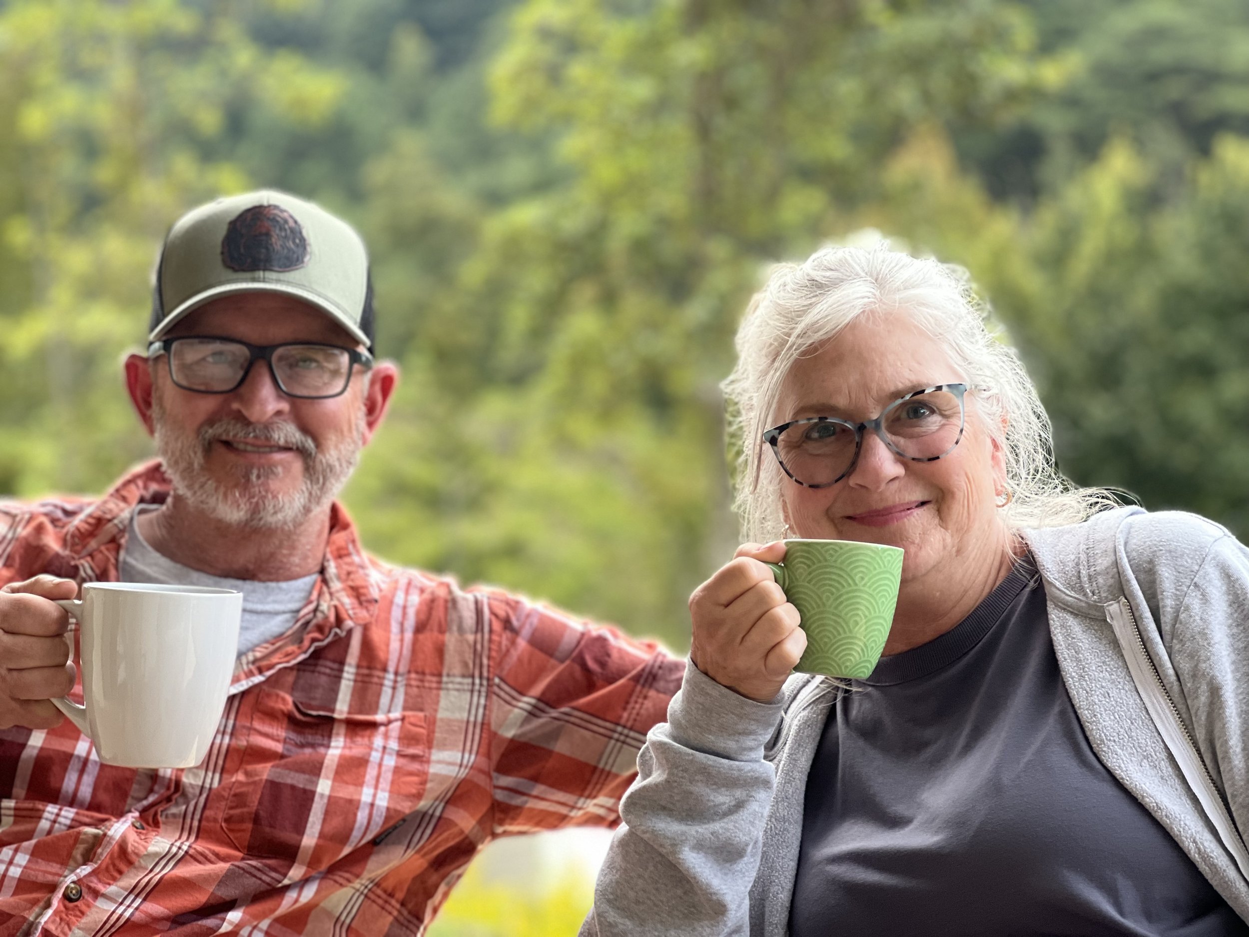 Smiling elderly man with glasses and a baseball cap, and smiling elderly woman with glasses, both holding coffee mugs and outdoors in a green, wooded area.