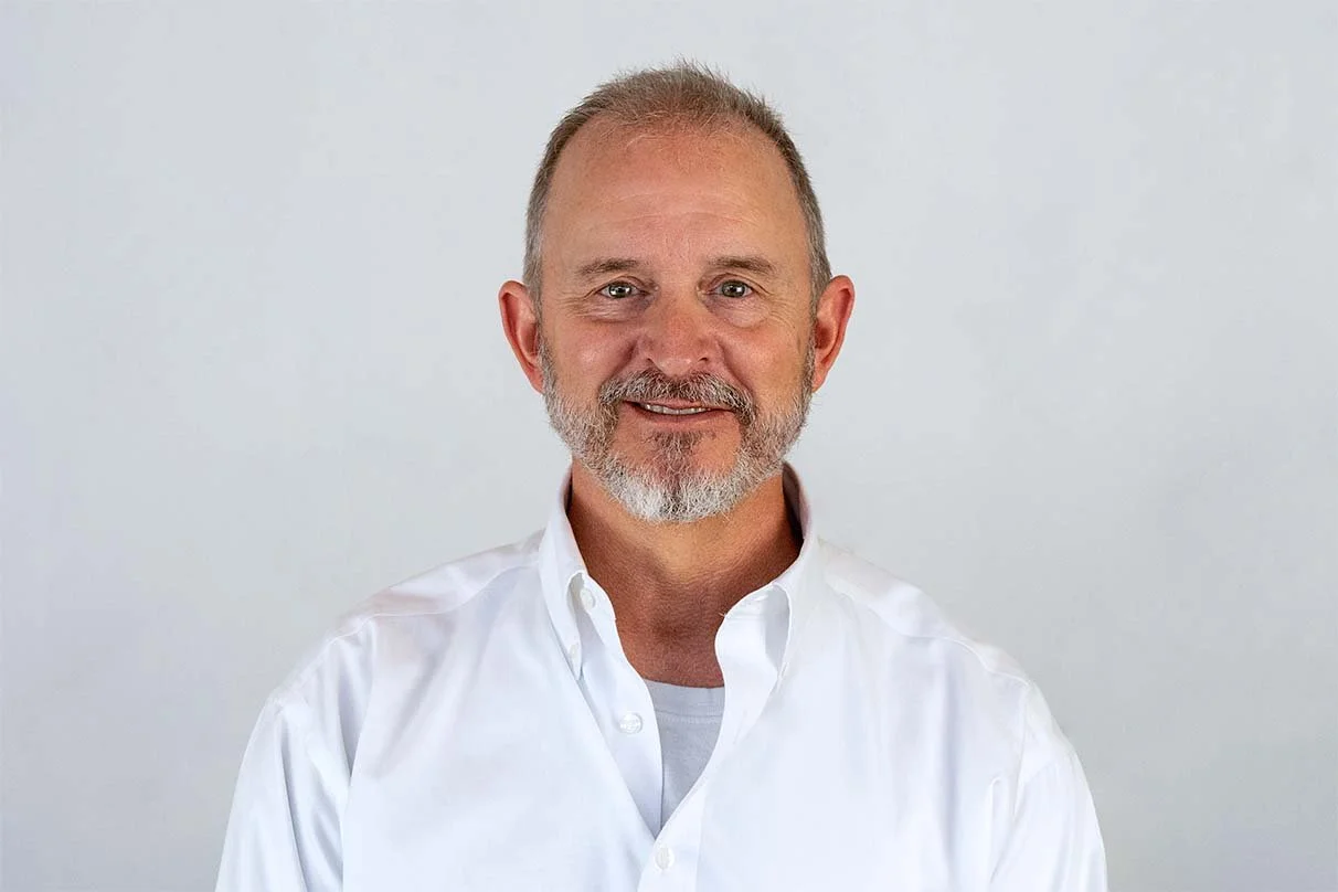 Smiling middle-aged man with a beard and short hair, wearing a white button-up shirt, against a plain light background.