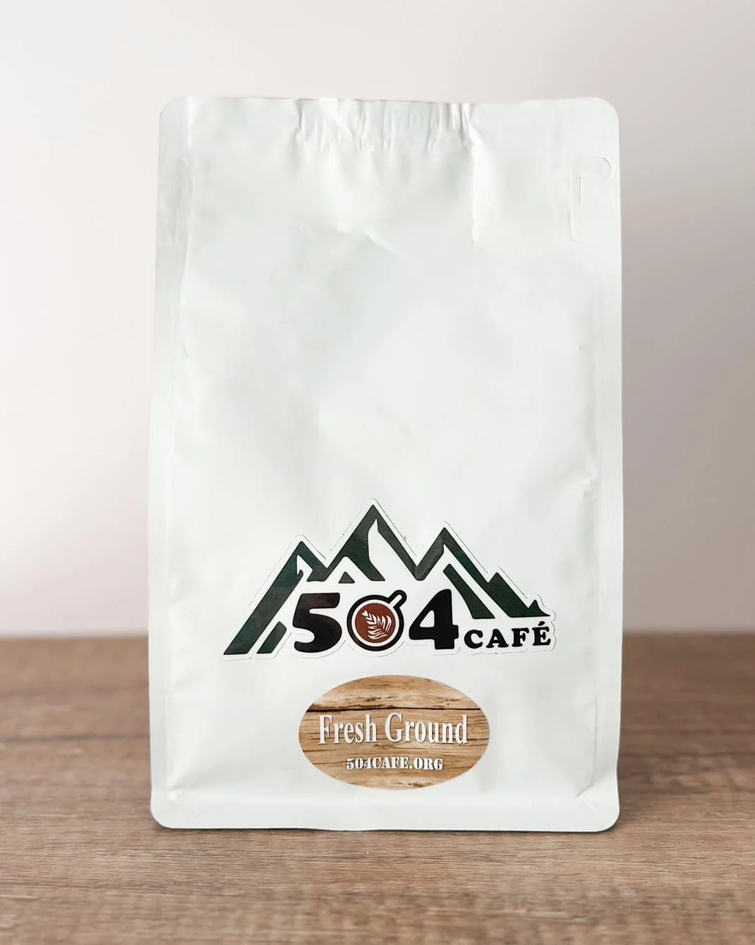 A white bag of coffee from 504 Café with mountain graphics and a label that reads 'Fresh Ground' and the website 504cafe.org.