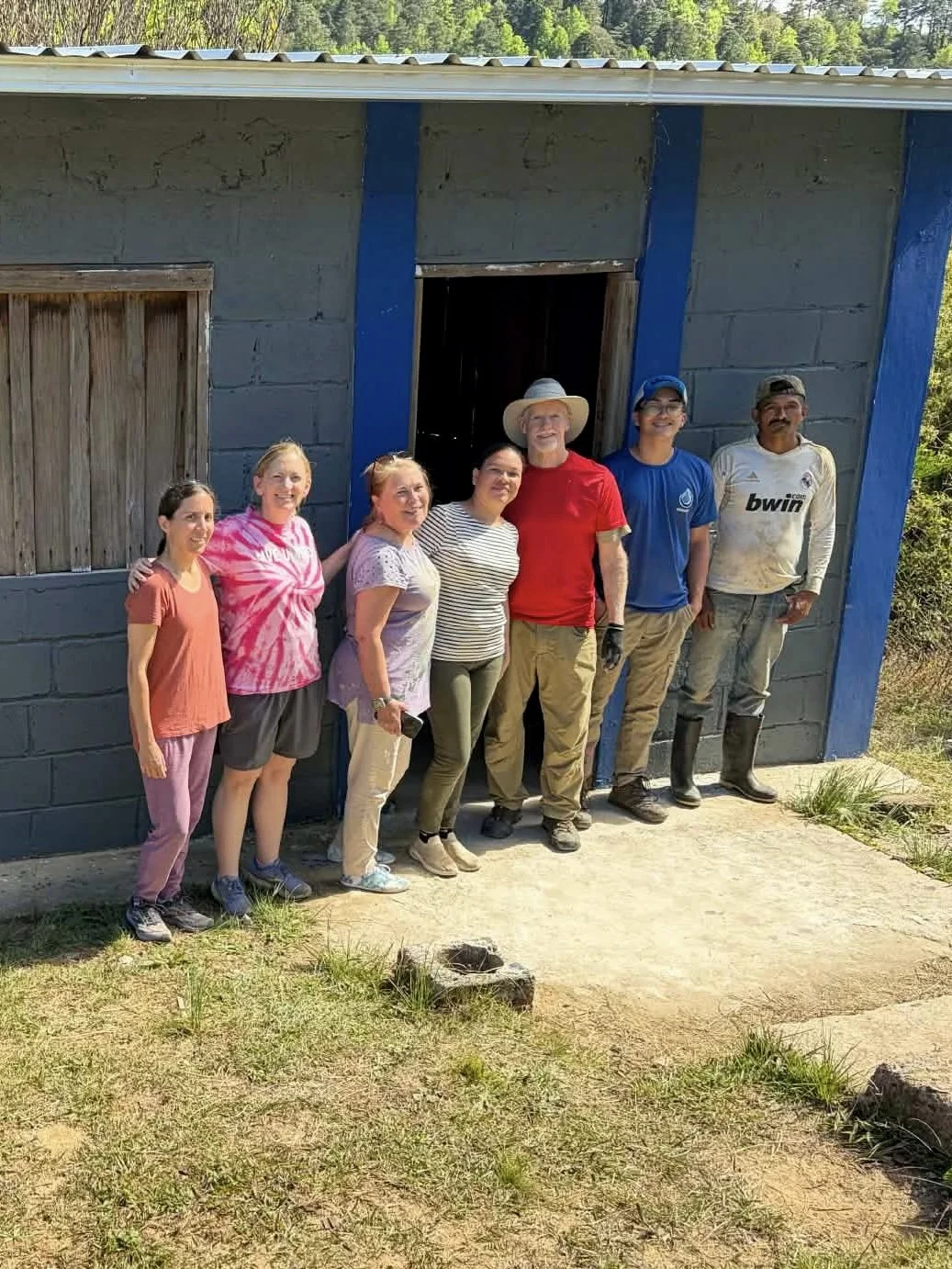 Group of seven people standing outside a small building with a gray cinder block wall and blue trim. They are smiling, and some are wearing work gloves and boots, suggesting they are involved in a building or renovation project.