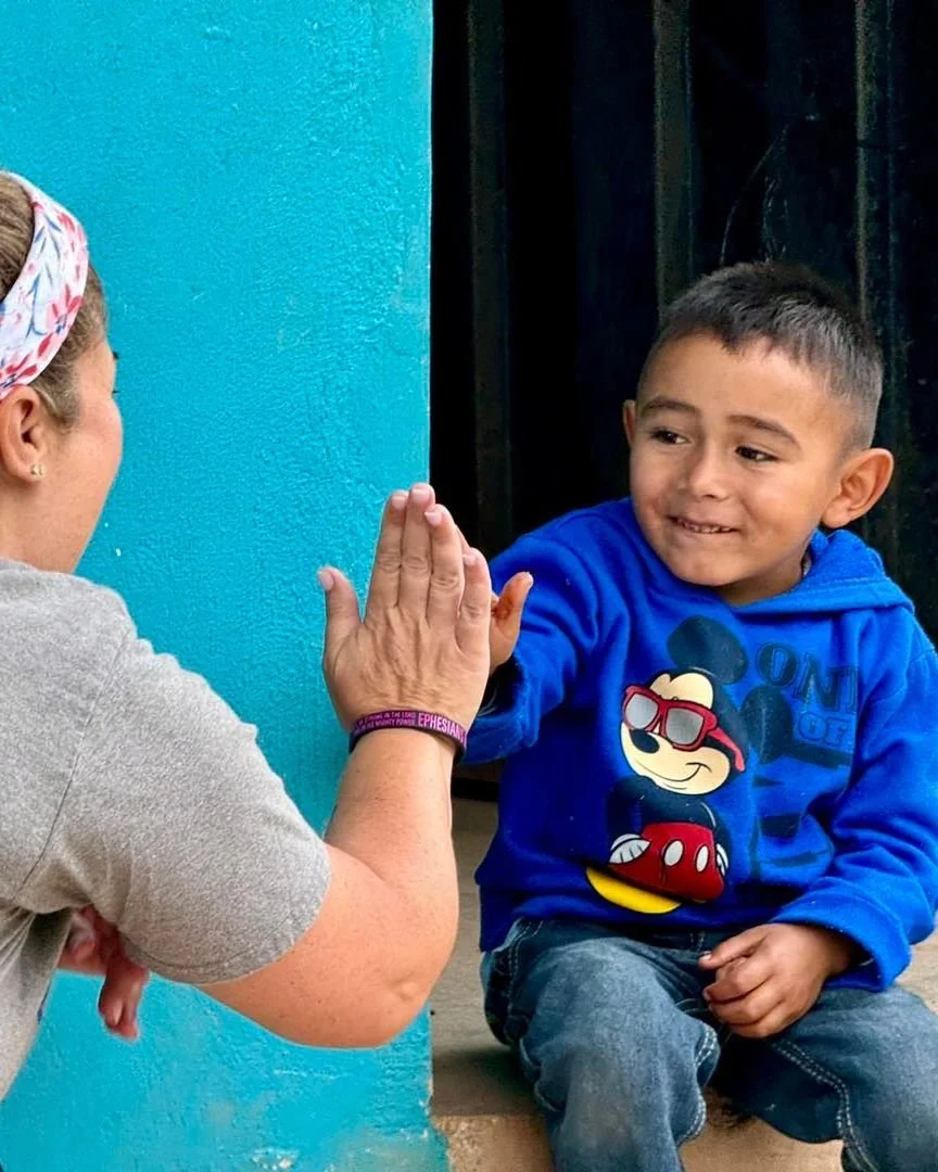 A young boy sitting on the ground giving a high-five to a woman facing him. The boy is wearing a blue hoodie with a Mickey Mouse print, and the woman has a headscarf and is wearing a gray shirt.