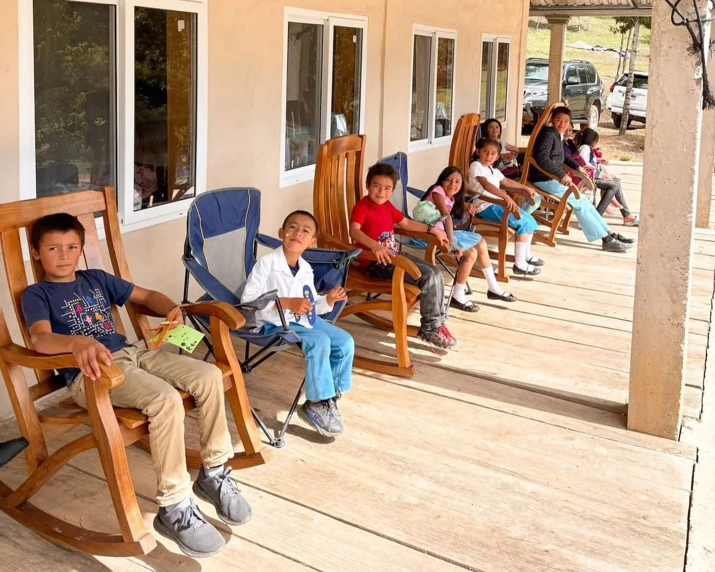 A group of children sitting on rocking chairs on a porch, smiling and looking at the camera.