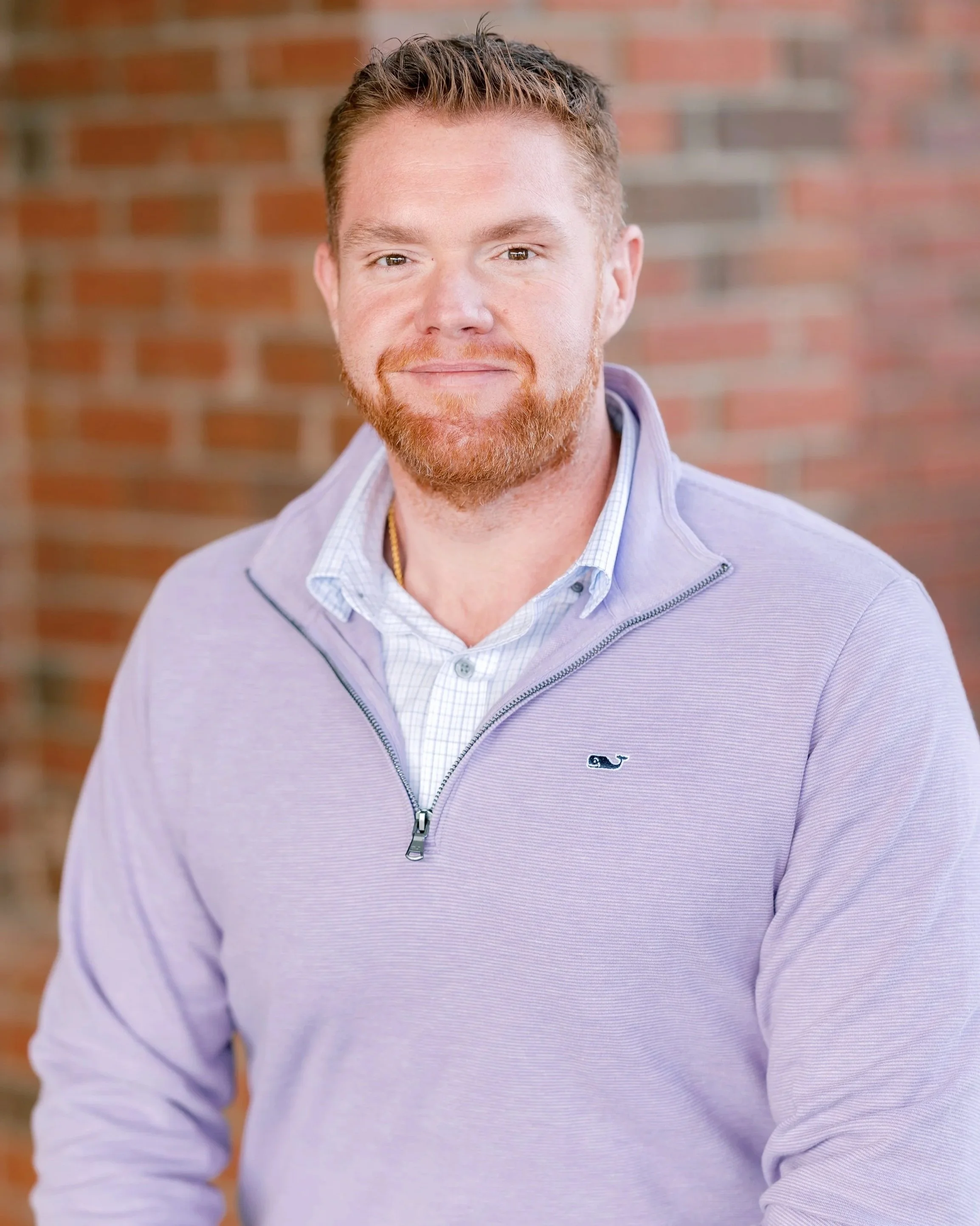 Close-up of a young man with red hair and beard, wearing a light purple quarter-zip sweater over a collared shirt, standing in front of a brick wall.