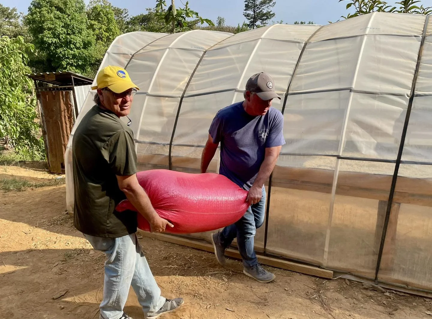 Two men are working together outdoors to move a large red bag, with one man holding the bag and the other helping.