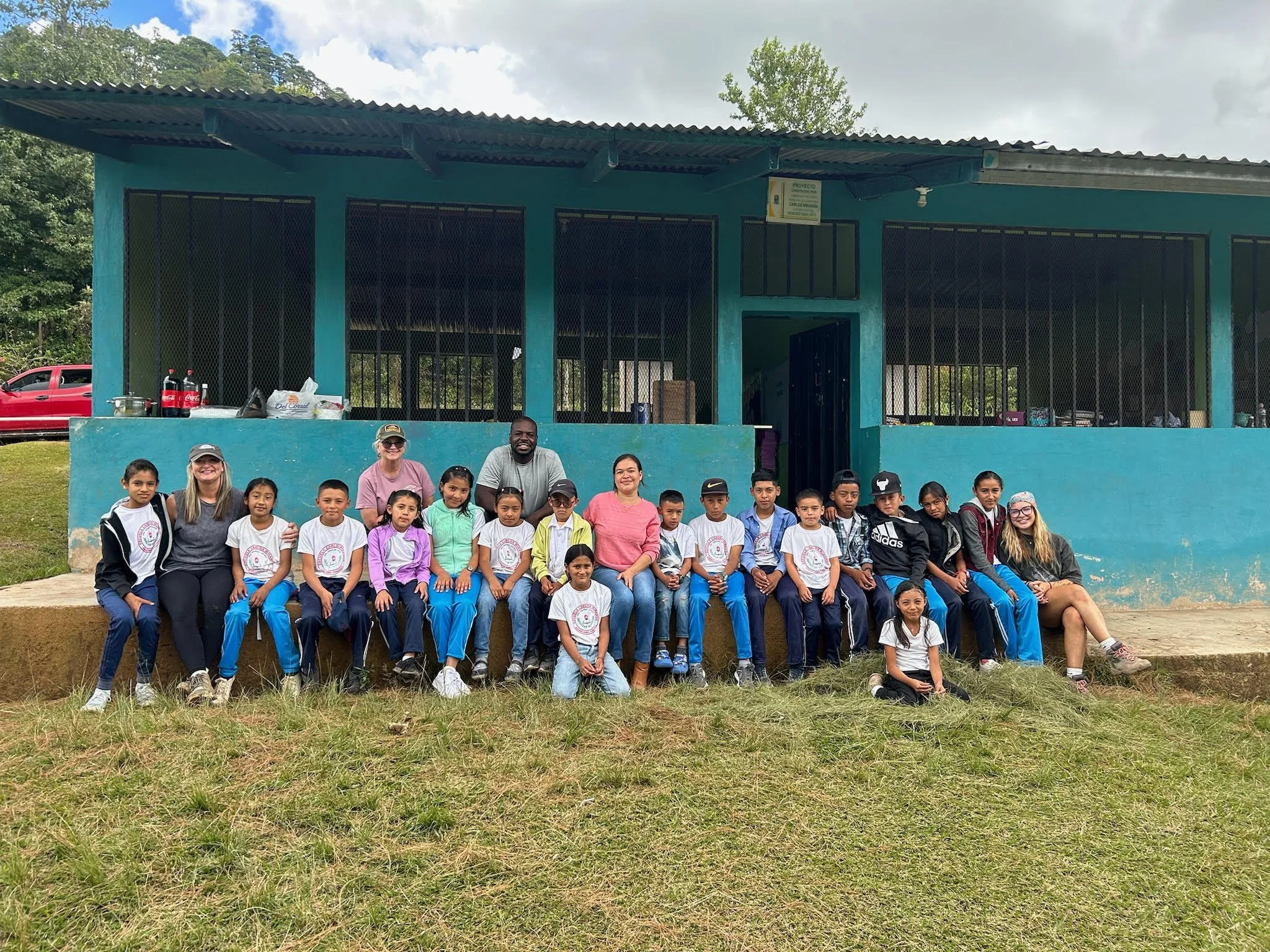 Group photo of children and adults outdoors in front of a blue building with a group of people smiling and posing.