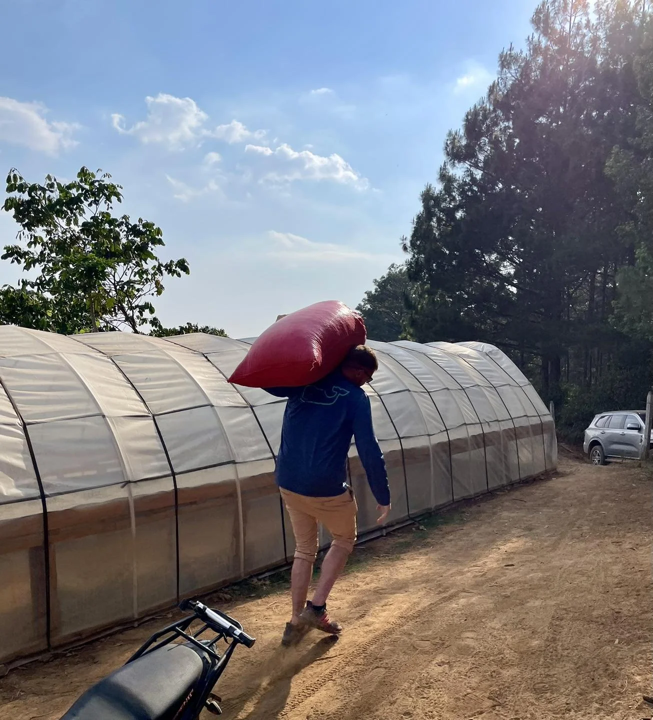 A person walking along a dirt path carrying a red bag on their shoulder. They are near a greenhouse structure with trees and a car in the background on a sunny day with a partly cloudy sky.