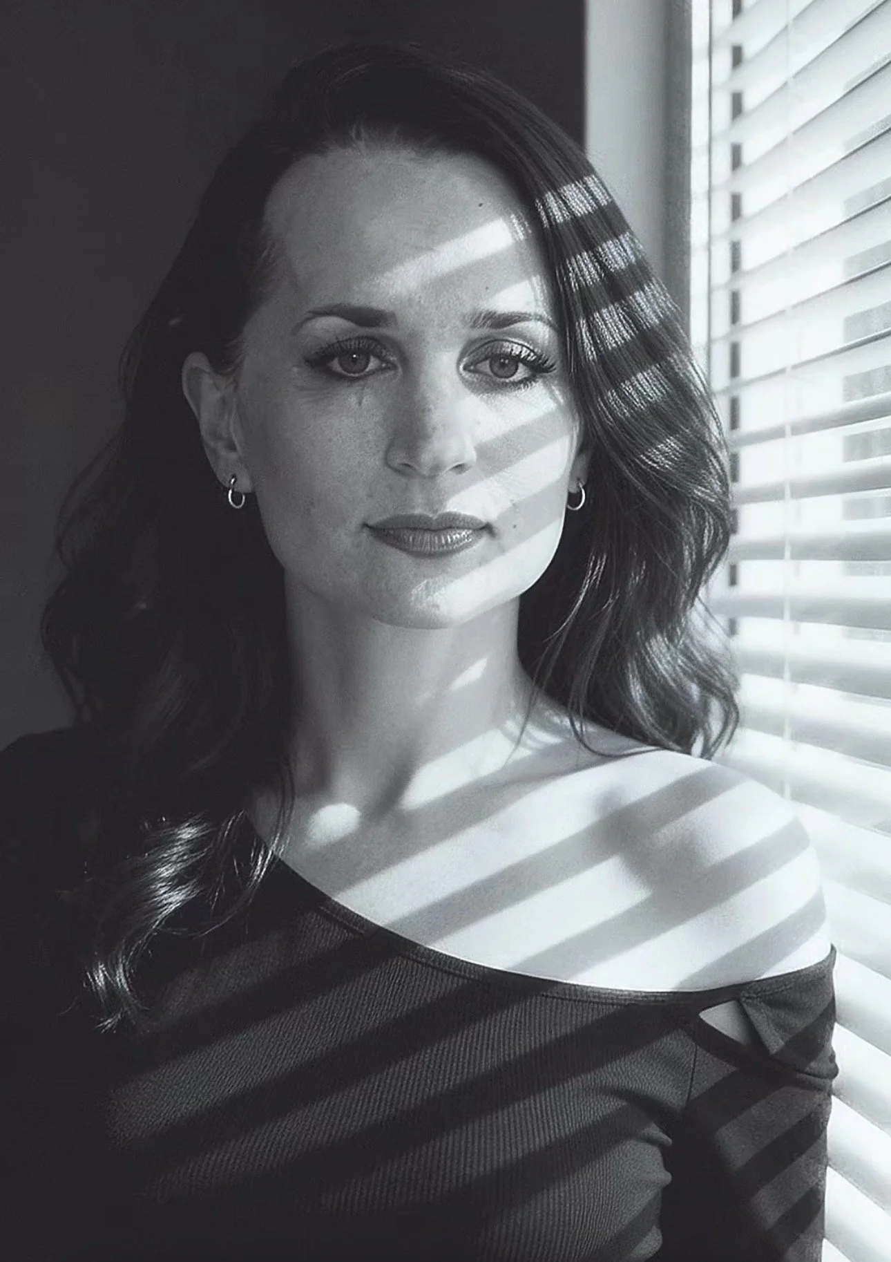 Black and white photo of a woman with wavy hair and hoop earrings looking at the camera, with sunlight and window blinds creating striped shadows on her face and clothing.