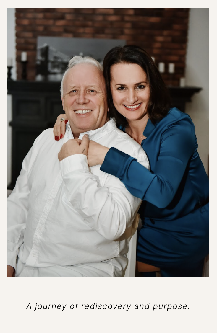 A smiling elderly man and a smiling woman hugging each other in a cozy living room with a brick wall and fireplace in the background.