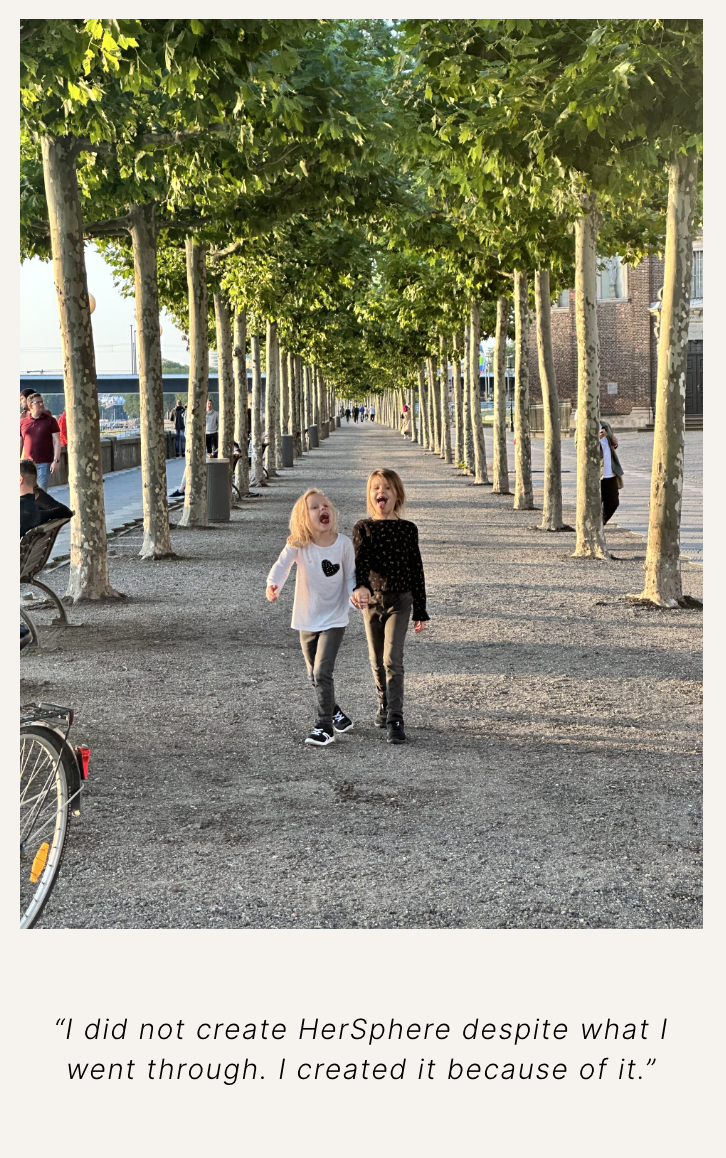 Two young girls laughing and walking hand-in-hand down a tree-lined pathway in a park or city setting during daytime.