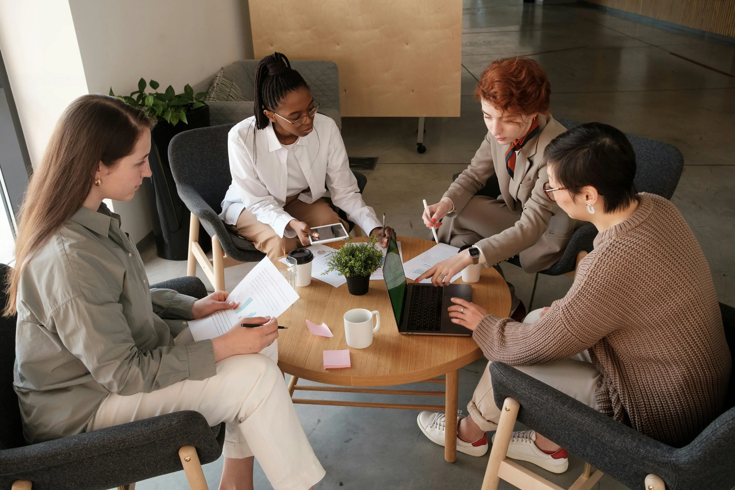 Four women sitting around a small round table, engaged in a discussion, with documents, laptops, and a small plant on the table in a modern office setting.