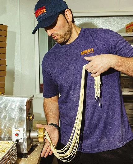 Chef hand-stretching fresh pasta dough beside a pasta machine in a restaurant kitchen.