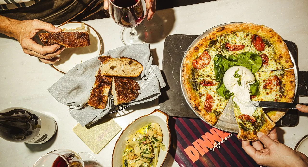 Guests sharing Roman-style Italian dishes, including pizza, handmade pasta, and wine, around a communal dining table.