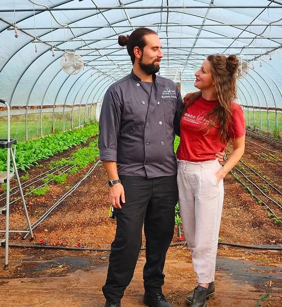Chef and local farmer standing together inside a greenhouse surrounded by growing produce.