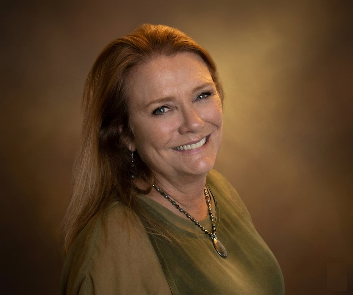 Portrait of a smiling woman with light skin and red hair, wearing a green top and a beaded necklace, against a brown gradient background.