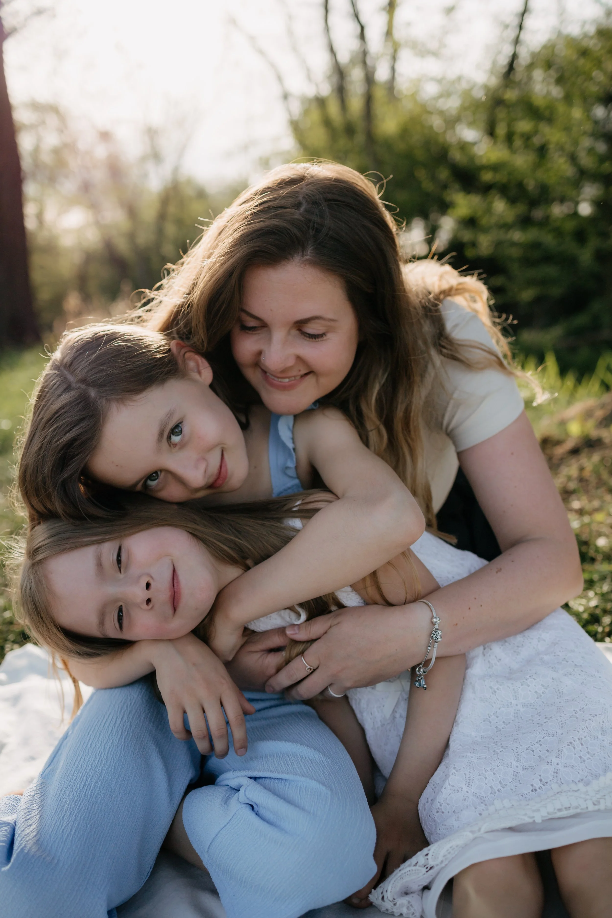 A woman with long brown hair and three young girls lying on the grass outdoors, hugging and smiling, with trees and sunlight in the background.