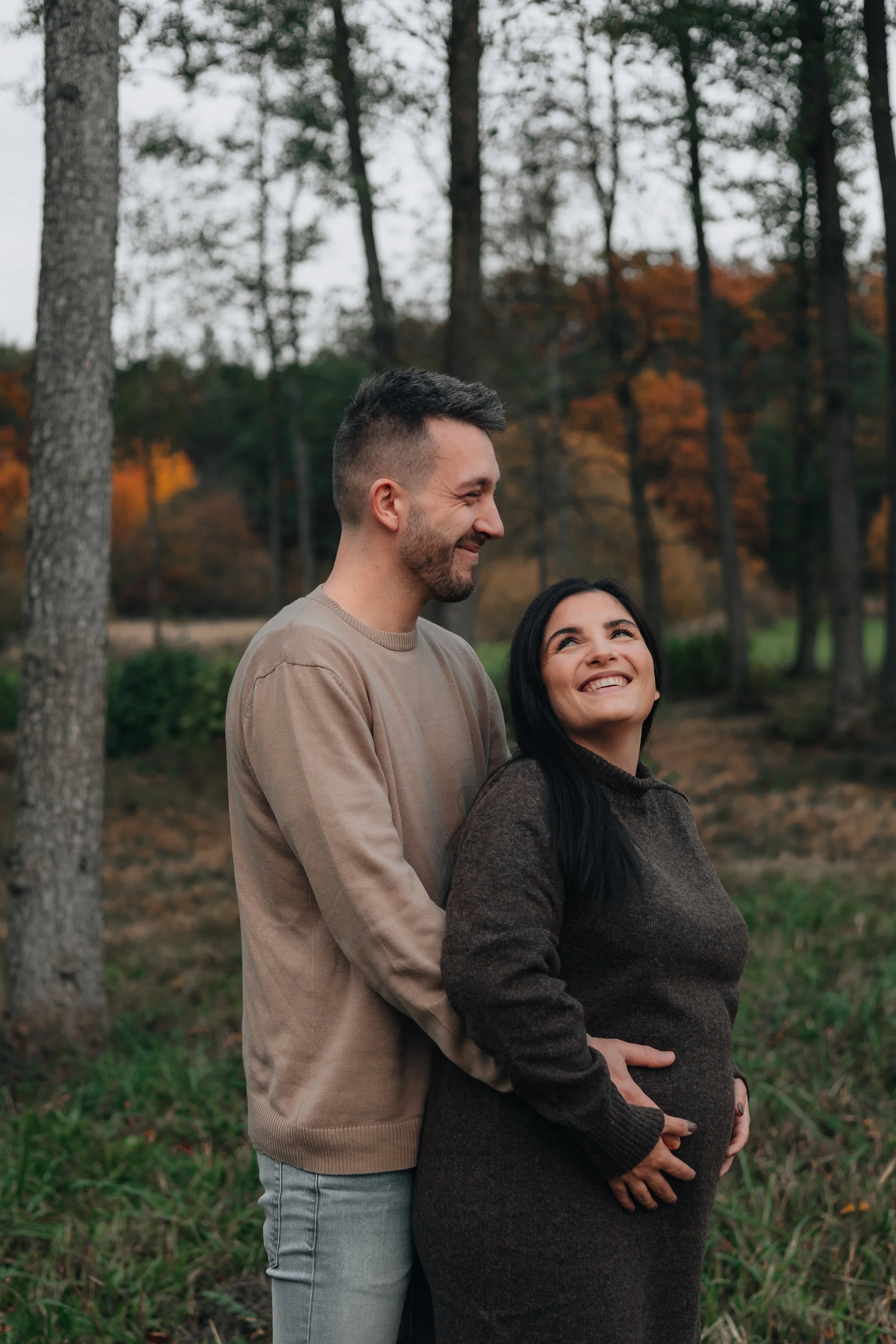 A man and pregnant woman smiling outdoors among trees during fall.