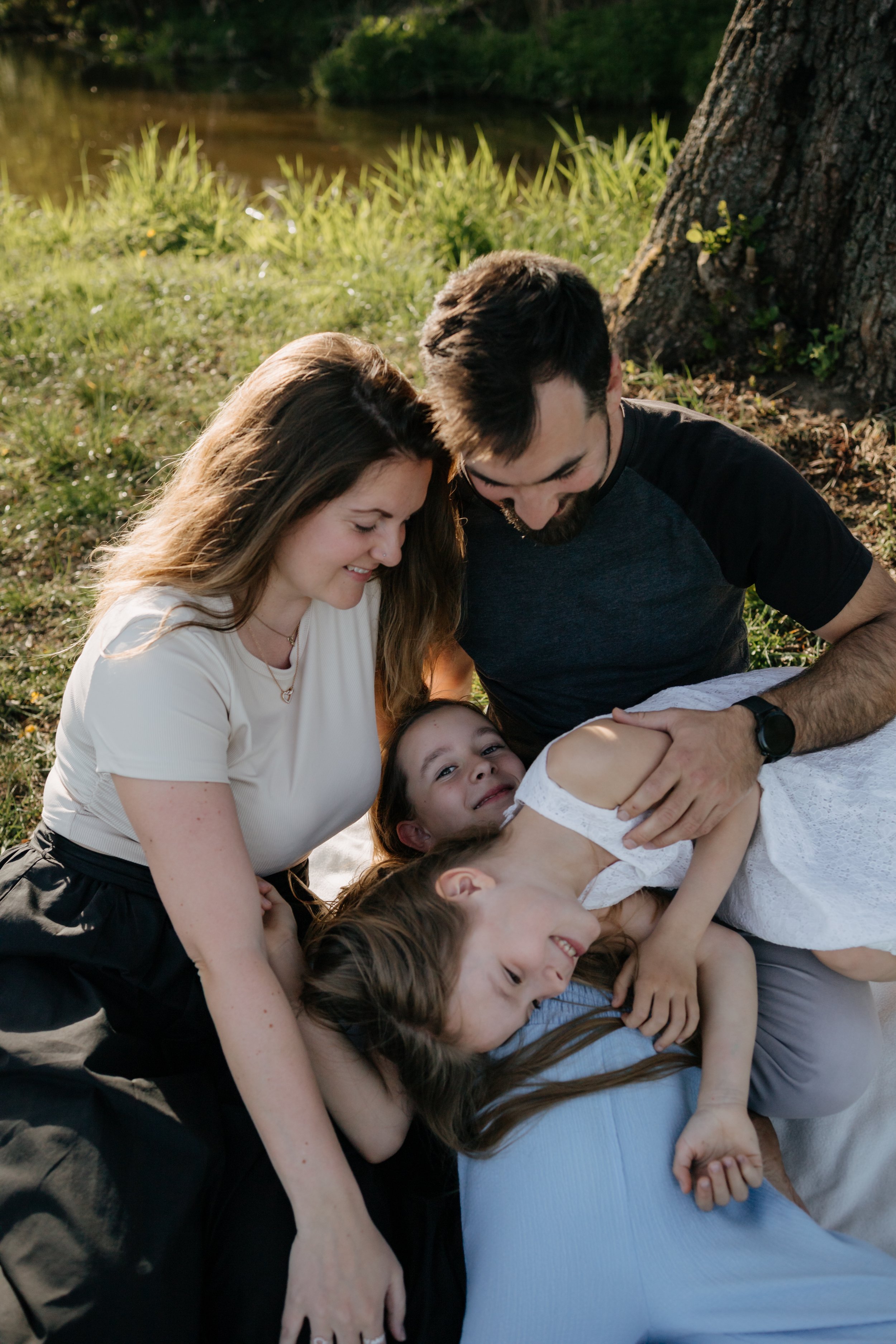 A group of people lying on the grass and huddled close together outdoors near a tree and a body of water, enjoying a sunny day.