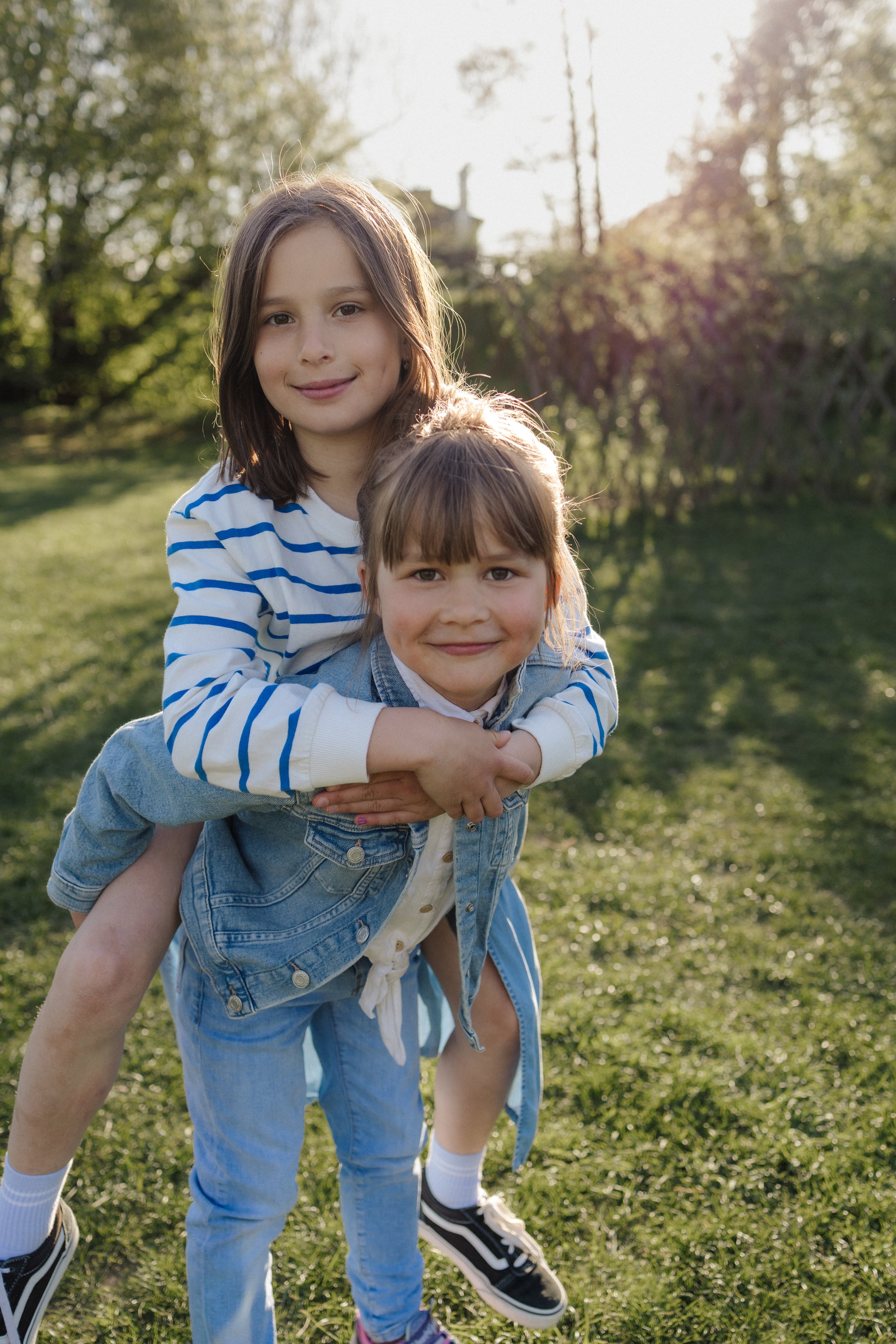 Two young girls, one with brown hair and the other with blonde hair, smiling and playing outdoors on a grassy field with trees in the background, bathed in late afternoon sunlight.