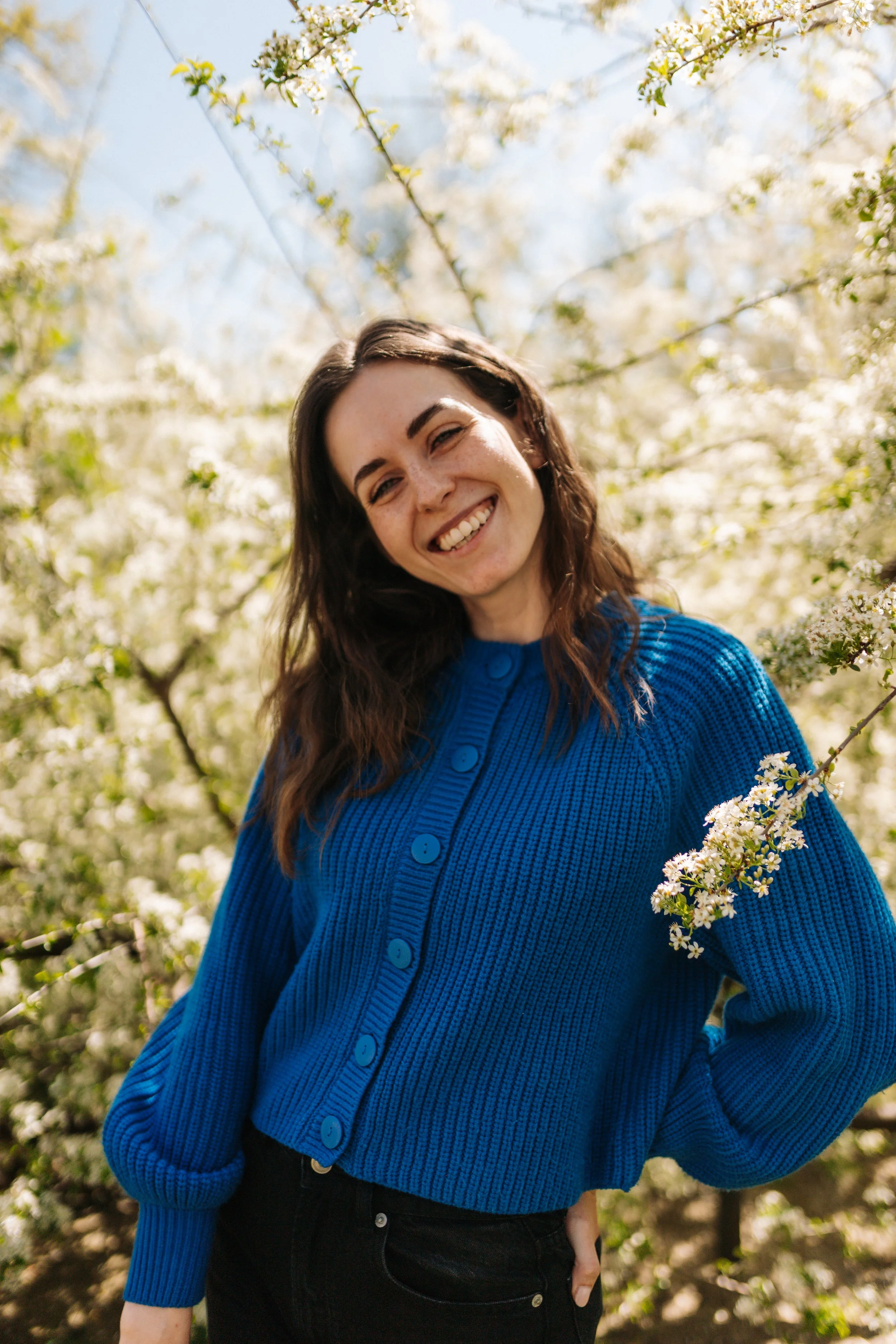 A young woman with long brown hair smiling, wearing a bright blue sweater with large buttons, standing outdoors among blooming white flowers and green foliage.