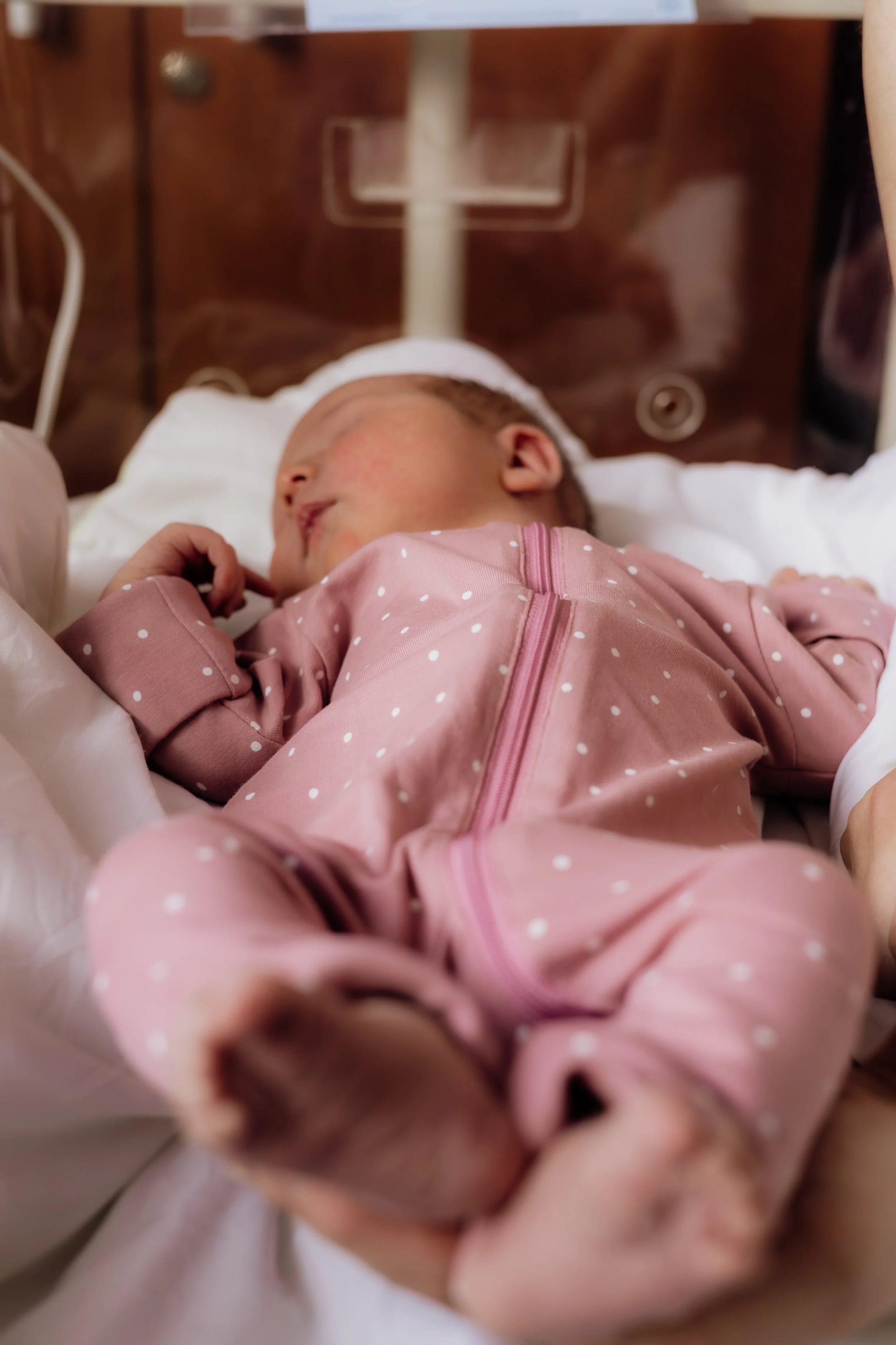 A baby lying in a hospital bed, wearing a pink polka dot onesie and a white hat, with medical equipment and a dark polished wooden headboard in the background.