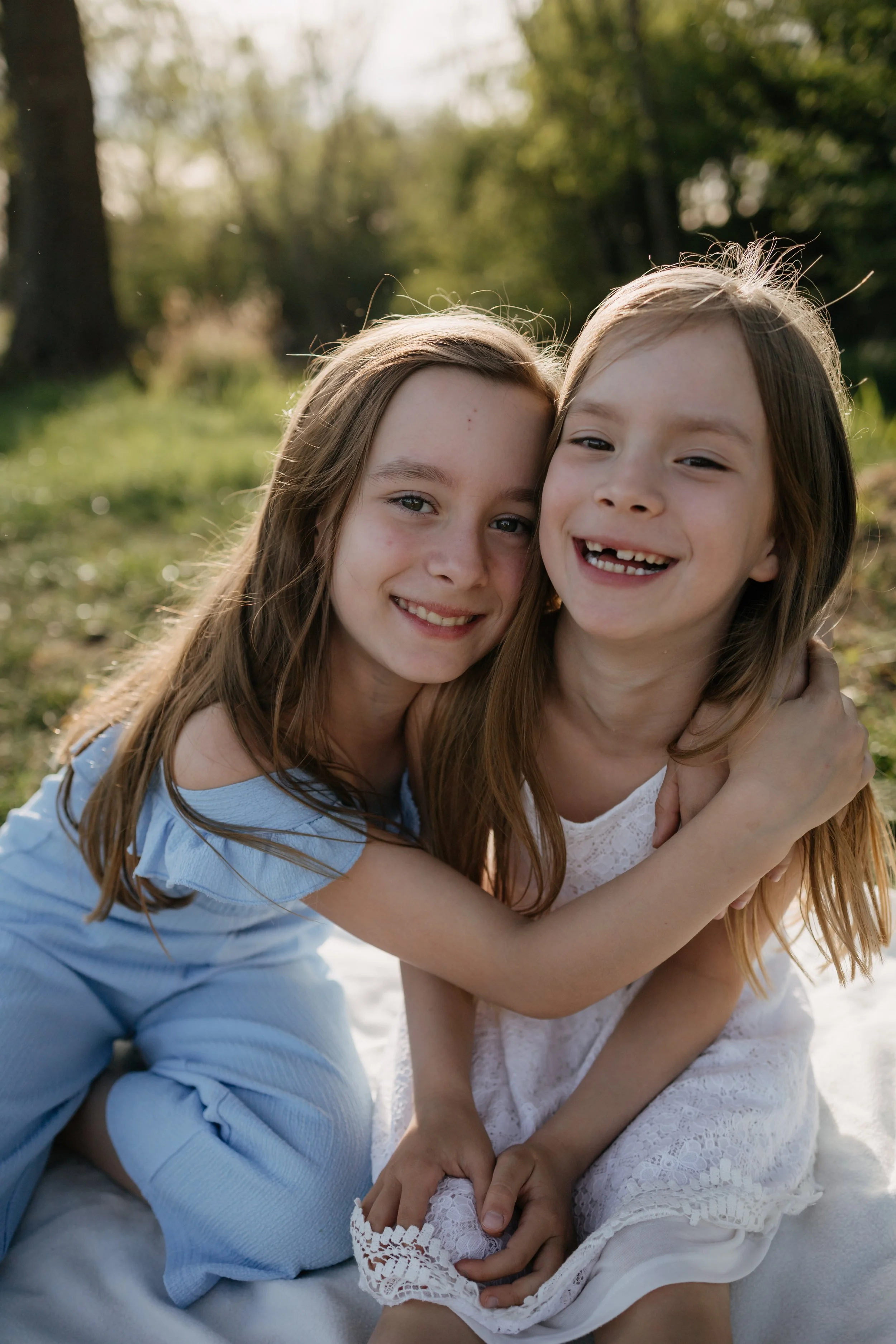 Two young girls hugging and smiling outdoors on a sunny day with trees and grass in the background.