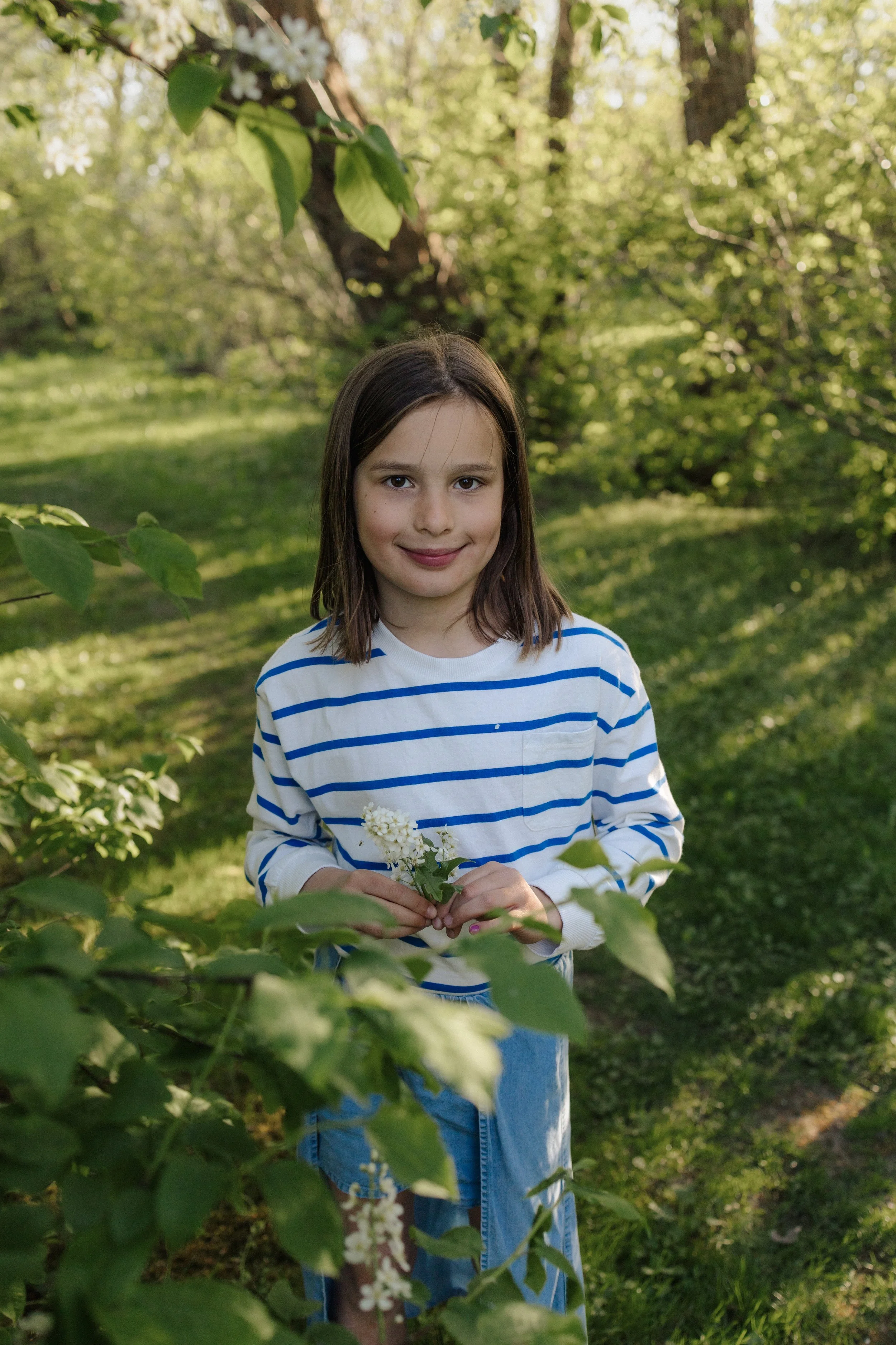 A young girl with long brown hair wearing a white and blue striped shirt and blue jeans, holding a small bouquet of white flowers, standing outdoors among green trees and foliage, smiling at the camera in a park or garden setting.