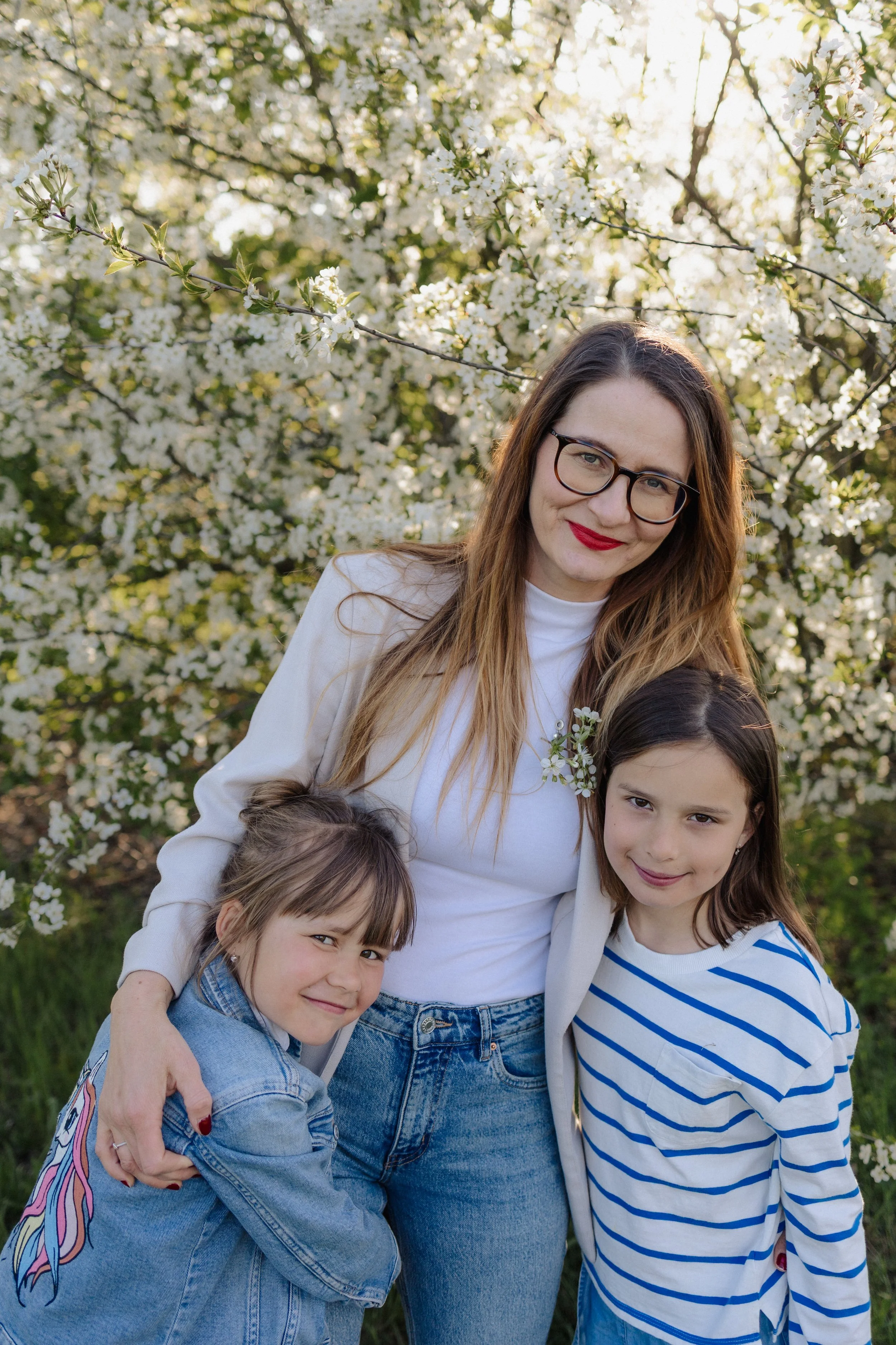 A woman with glasses and red lipstick standing among four children in front of white flowering trees during the daytime.