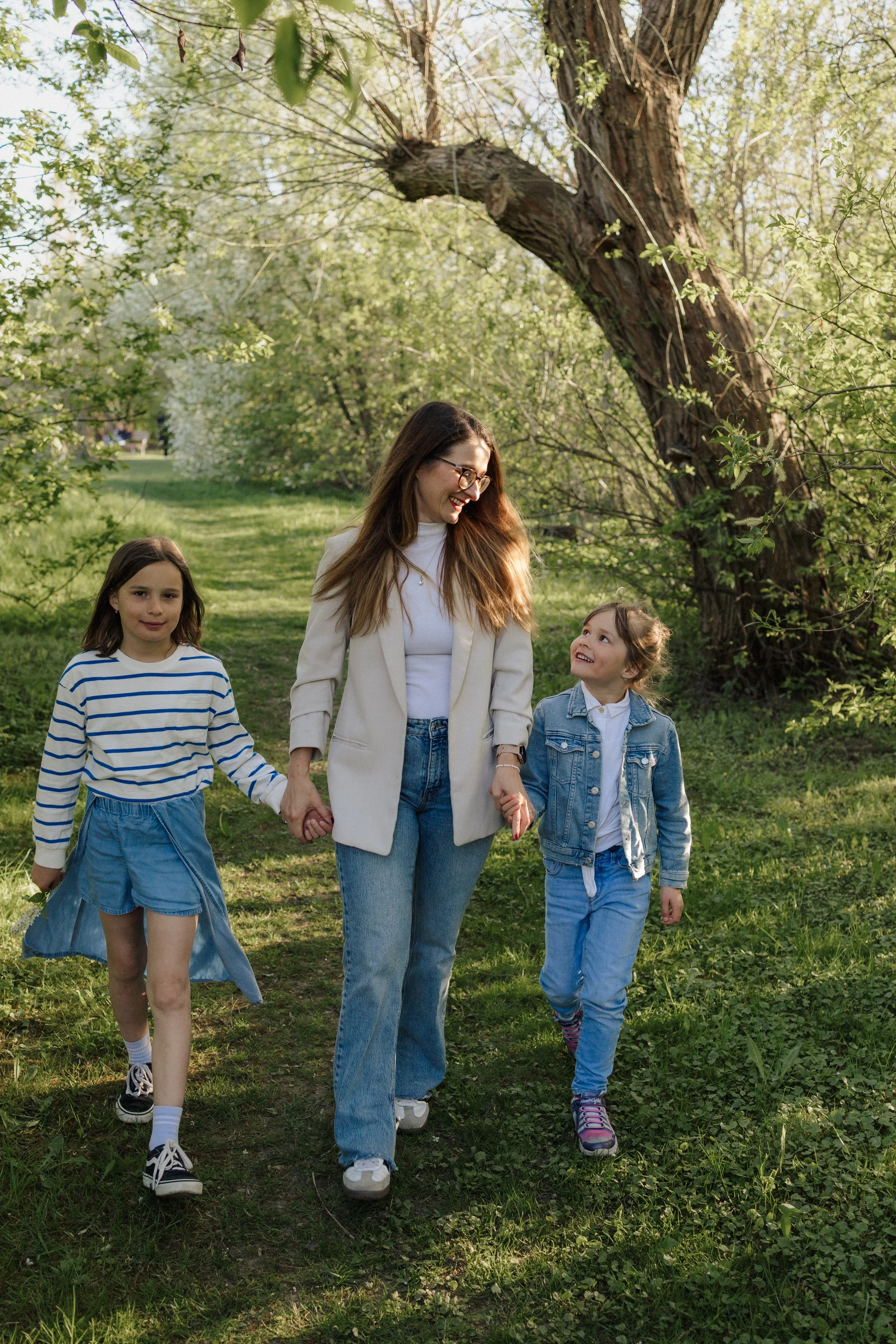 A woman and two young girls walking hand-in-hand through a park or forested area with green grass, trees, and sunlight. The woman has long brown hair, glasses, and is wearing a white blazer and blue jeans. The girl on the left is wearing a blue and w